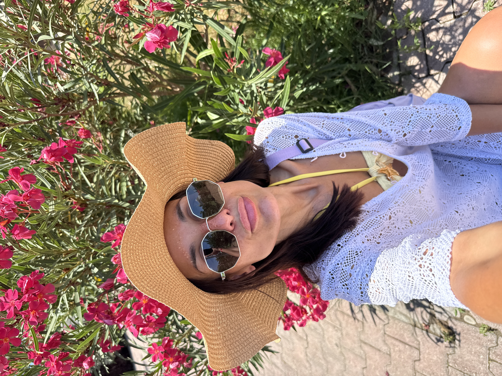 A woman wearing a straw hat and sunglasses poses for a selfie in front of a bush with pink flowers. She is wearing a white lacy cover-up over a yellow bikini top and a light purple bag strap is visible. The background is a mix of greenery and pink flowers, with a paved walkway and sunlight casting shadows. The setting is outdoors in a sunny environment.
