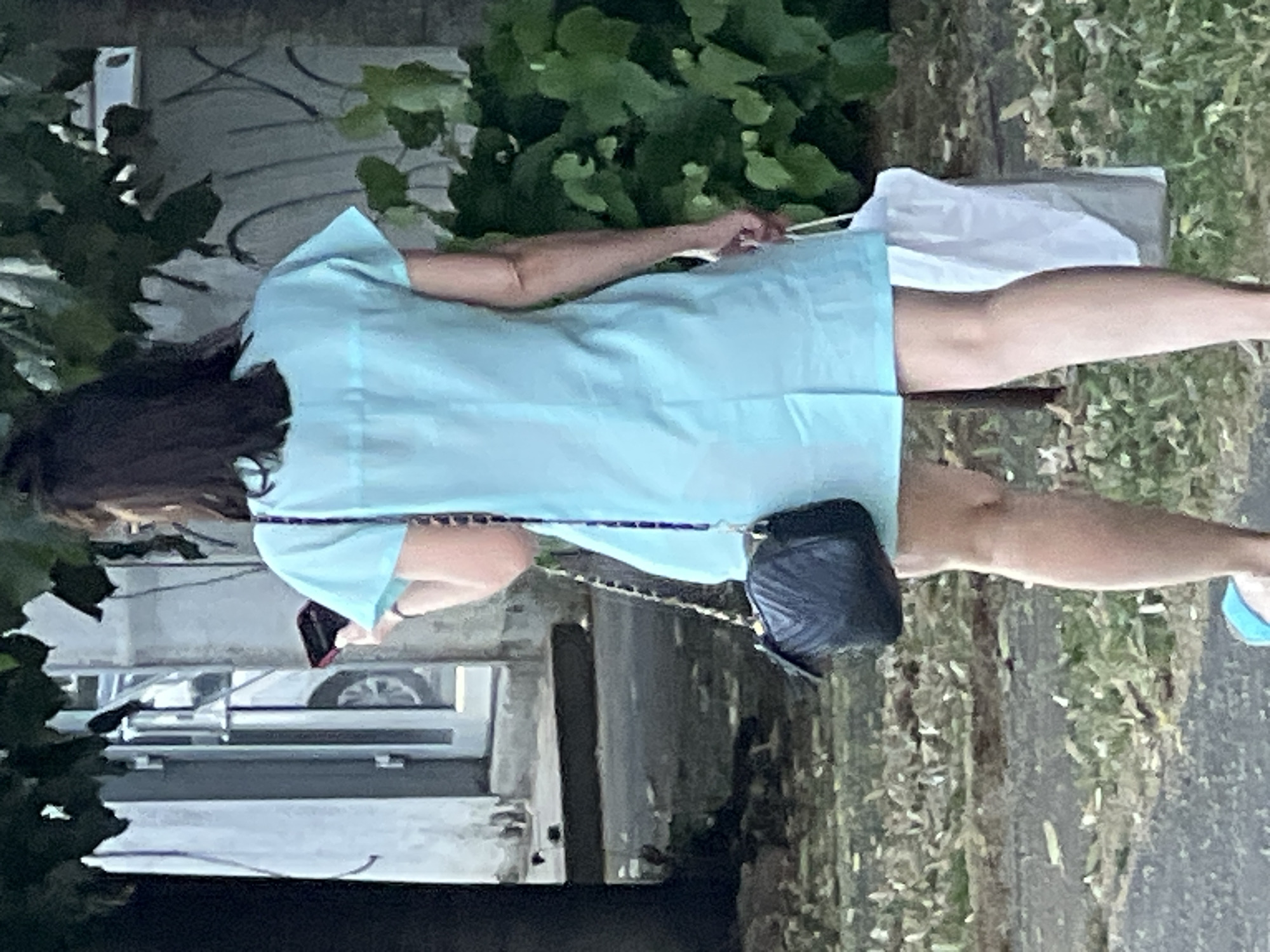 A woman is walking away from the camera, holding a dark-colored, quilted purse with a gold chain strap on her left shoulder. She is wearing a light blue, short-sleeved dress and carrying a white bag in her right hand. The woman is holding a dark-colored smartphone in her left hand, which is visible near her shoulder. She is walking on a paved surface with scattered leaves. In the background, there is foliage, a light-colored wall with some graffiti, and a glass door with a white car visible inside. The scene appears to be outdoors in a city setting. Based on the provided context, this image was taken in Krasnodar, Russia.