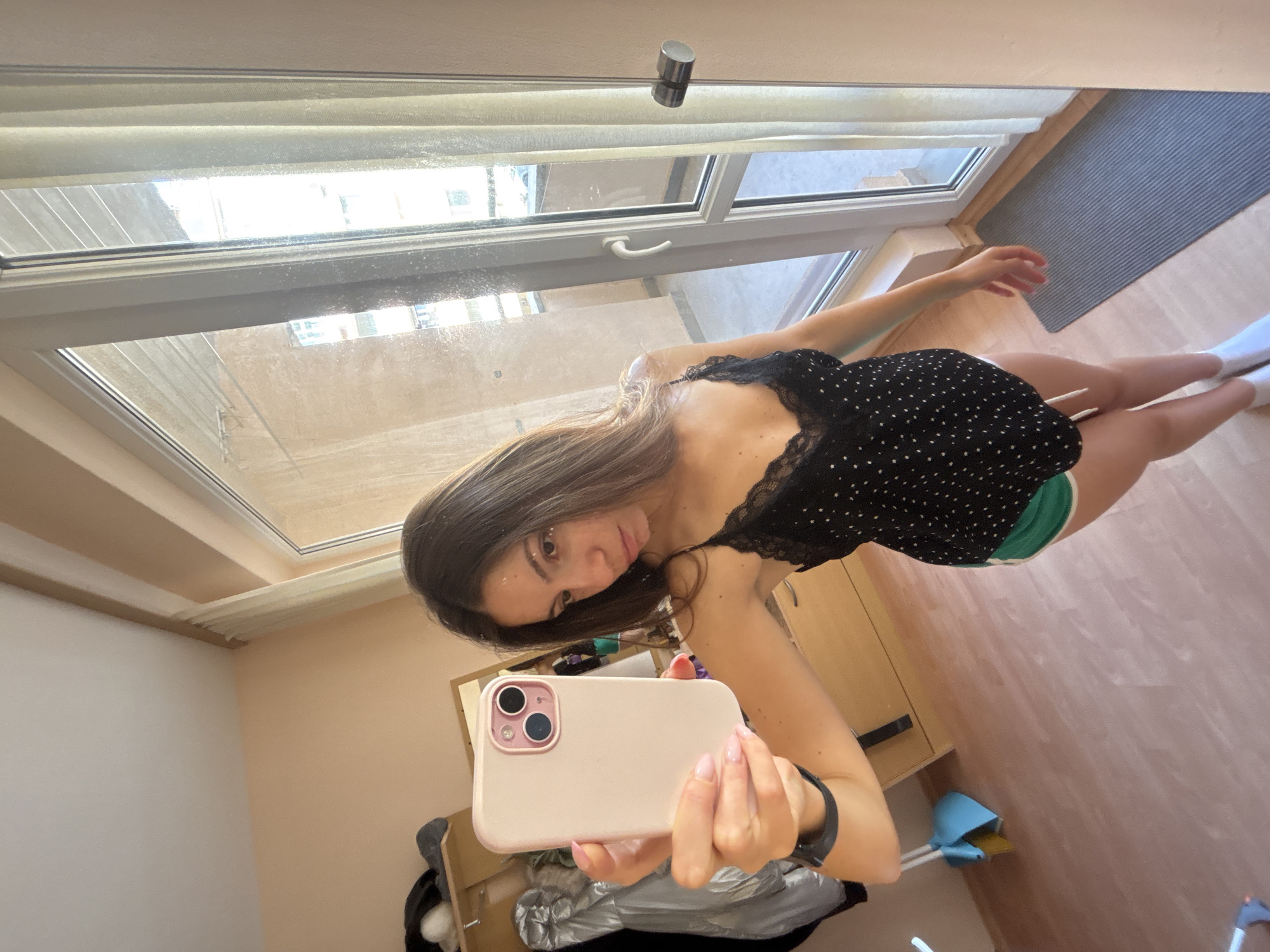A young woman with long brown hair takes a selfie in a room with wooden floors and peach-colored walls. She is wearing a black camisole with white polka dots and green shorts. In her right hand, she holds a light pink smartphone. To her right is a large window with sheer curtains and a sliding glass door that opens to a balcony. A gray yoga mat lies on the floor in front of the door. In the background, a wooden dresser and a rack with clothing are visible. The scene appears to be an indoor residential setting. Varna, Bulgaria is the city where this image was taken.