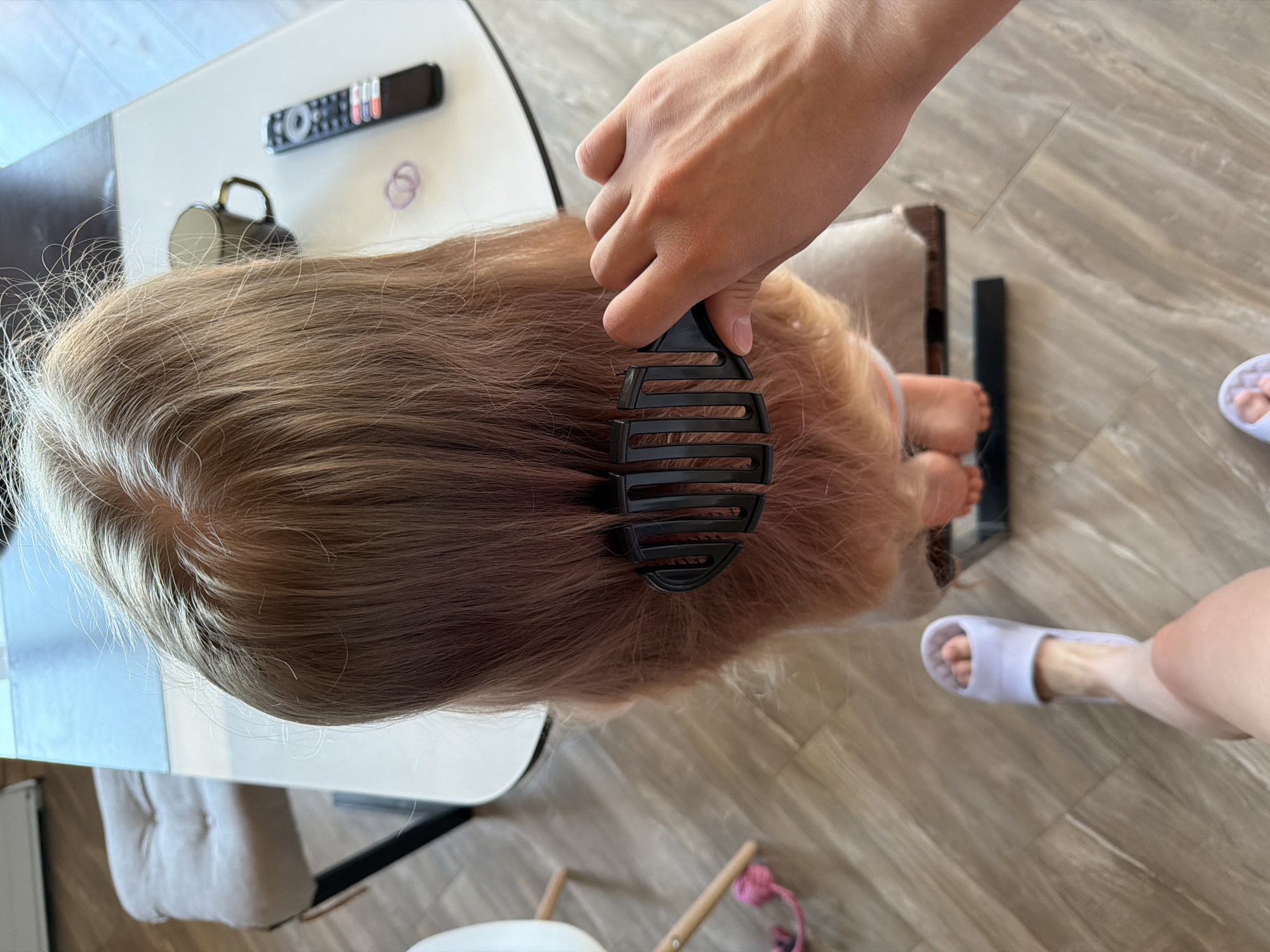 A top-down shot depicts a person with fair hair being groomed indoors. An adult hand holds a large, black comb and runs it through the child's hair. The child sits on a chair. Their feet are visible, one pair bare and another foot wearing a light-colored slipper.
The scene is set near a table with a white and black top, on which rest a black TV remote, a grey mug, and two hair bands. The floor is a light-colored wood-grain pattern. A pink hair tie is lying near the bottom of the frame.
The city and location are not discernible from the image.