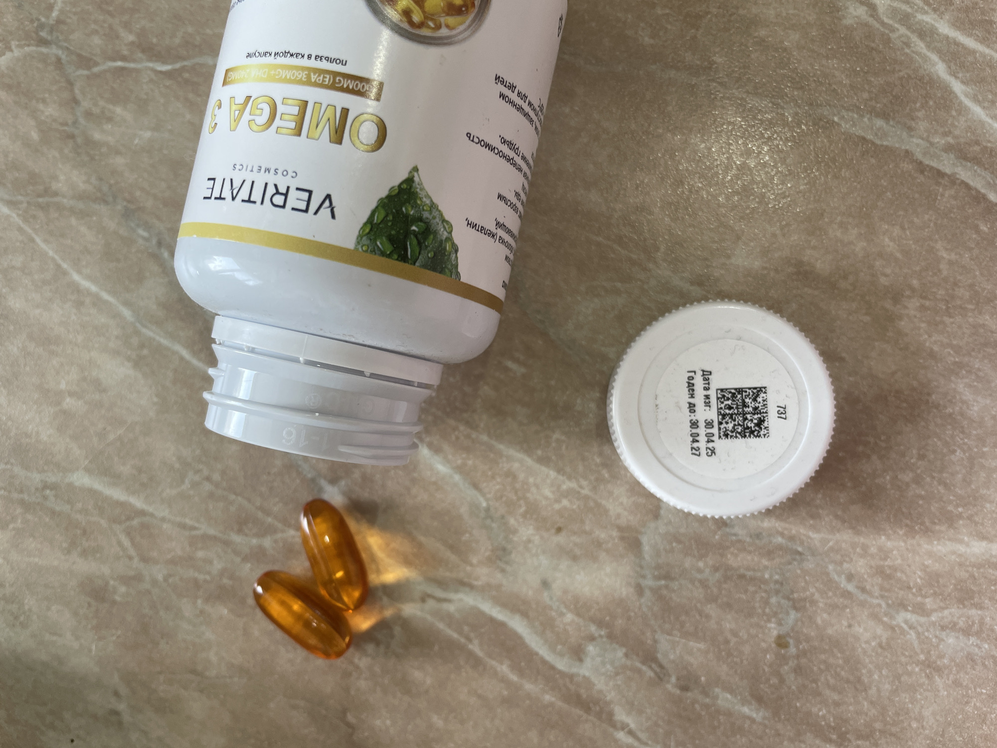 The image shows a bottle of "VERITATE COSMETICS OMEGA 3" pills with two capsules next to it. The bottle is white with text in Russian. The cap of the bottle is white with the date of manufacture as 30.04.25 and the expiration date as 30.04.27, also in Russian. The background is a light brown surface. The pills are golden in color. There is a marking "1-16" on the bottle. The location is unknown.