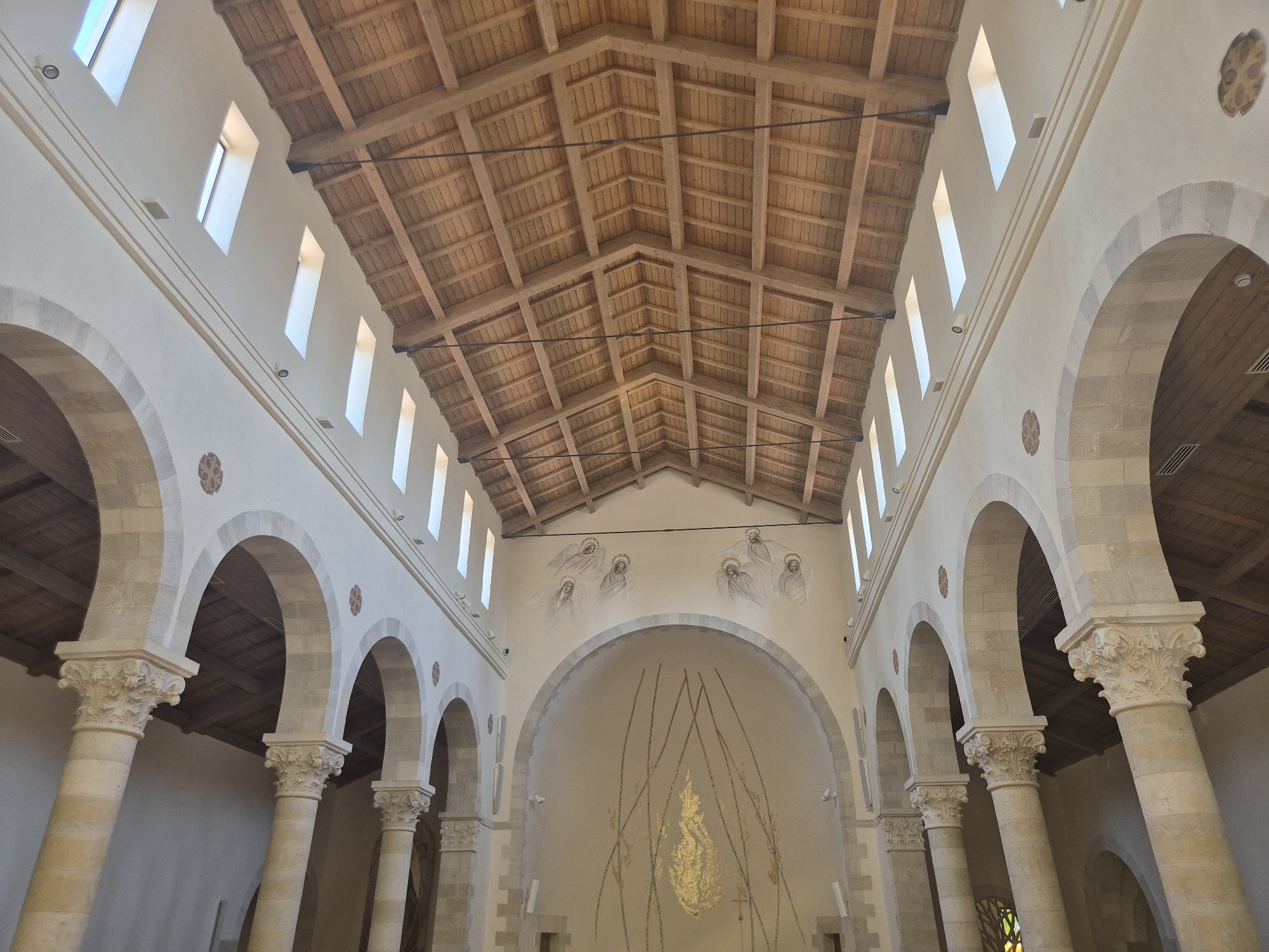 The image shows the interior of a church in Abu Gosh, Israel, featuring a high, wooden-beamed ceiling and stone arches supported by ornate columns. Sunlight streams in through tall, narrow windows positioned high on the walls. The far wall of the apse is decorated with a painting of angelic figures and a central golden, flame-like mosaic. The architecture is Romanesque in style, with rounded arches and carved capitals on the columns.