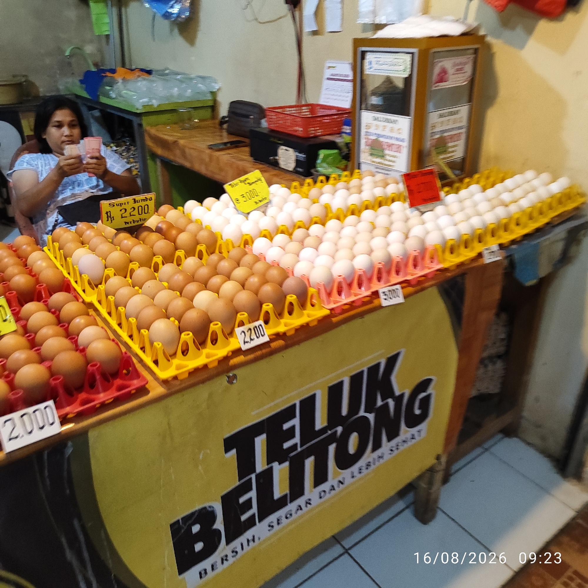 The image depicts an outdoor market stall in Pembaharuan, Indonesia, showcasing a variety of eggs for sale. A woman, presumably a vendor, sits in a chair on the left, counting cash. The stall is filled with rows of eggs arranged in yellow and red plastic trays. Several price tags are visible, indicating prices such as "2.200", "3.000-", and "5.000". A large yellow banner with black lettering reads "TELUK BELITONG BERSIH, SEGAR DAN LEBIH SEHAT", suggesting the brand or slogan associated with the egg vendor. In the background, a wooden counter holds various items, including a red basket, a dark box, and some packaged goods. The overall setting suggests a typical day at a local Indonesian market.
