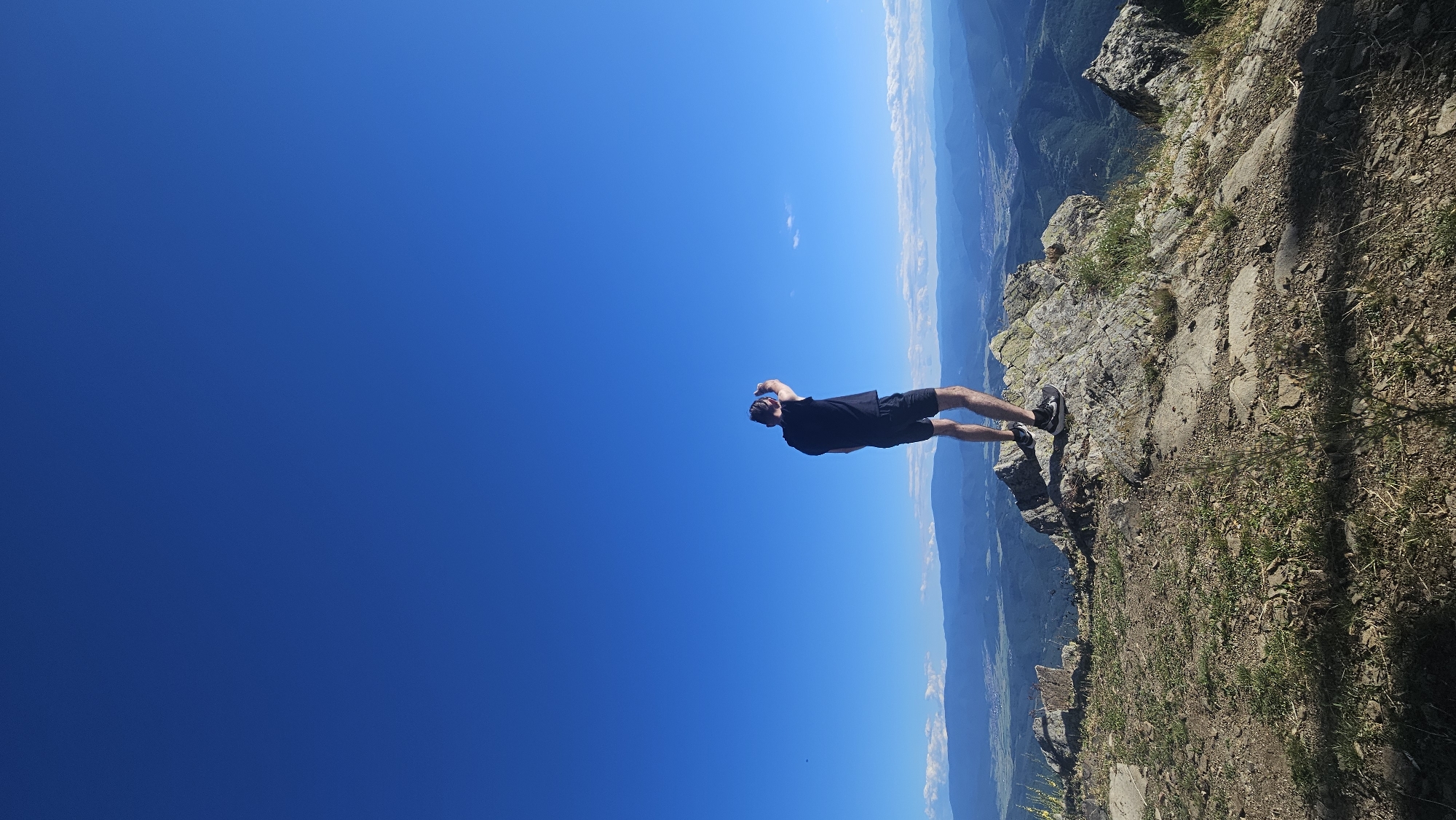 A man stands on a rocky outcropping, his back to the camera, looking out over a vast valley under a clear blue sky. He is wearing a black t-shirt and black shorts with athletic shoes. His hands are raised to his head, as if shielding his eyes from the sun or adjusting something. The foreground shows the rugged terrain of the outcropping with sparse vegetation and rocks. In the background, rolling hills and mountains recede into the distance, with a small town or village visible in the valley below. The overall scene suggests a moment of contemplation or awe at the expansive landscape. The location is identified as the city of Simeonovets, Bulgaria.