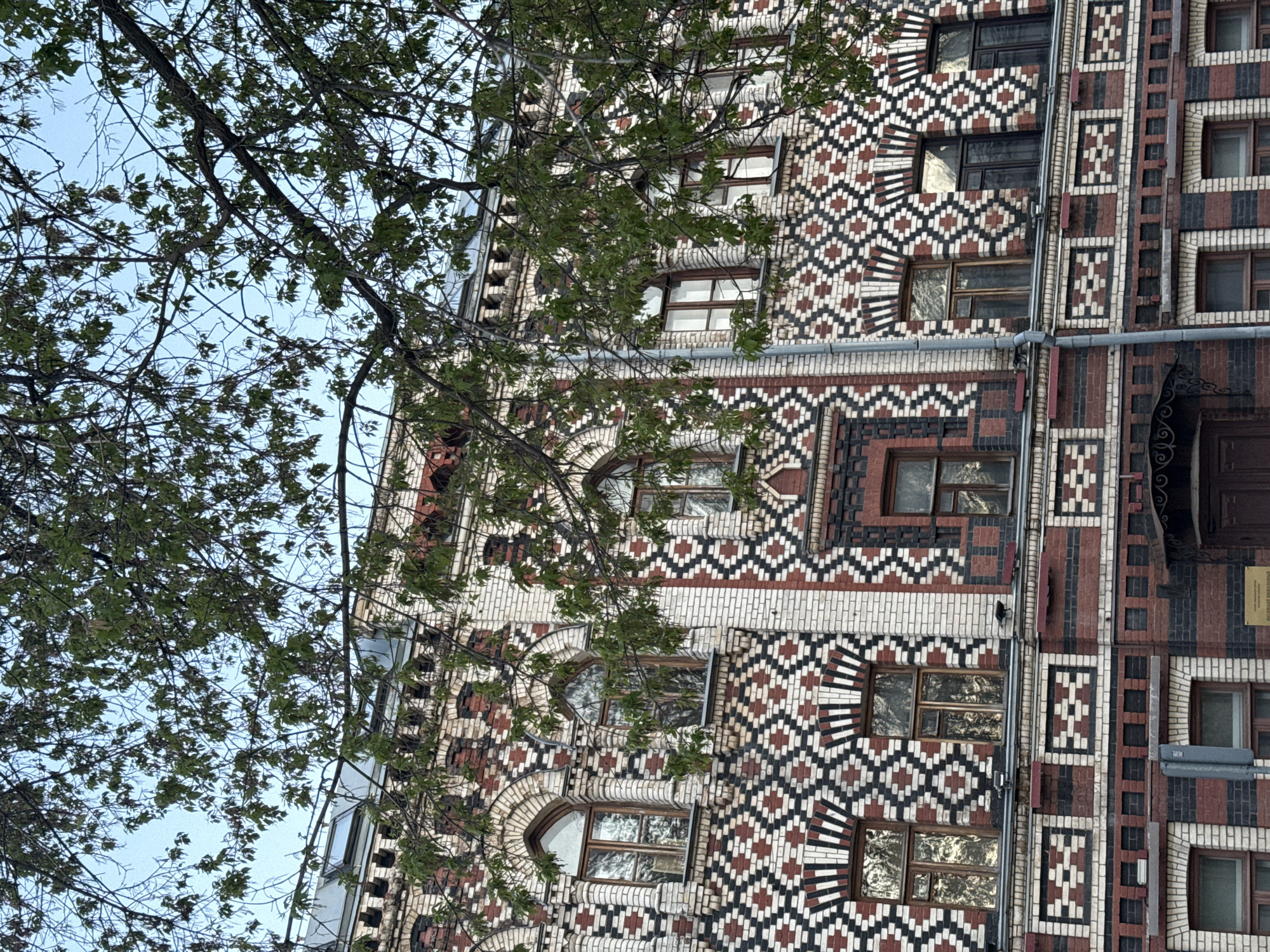 The image shows a building in Moscow, Russia, specifically the Igumnov House at Yakimanka Street, 43. The exterior of the building is characterized by its ornate brickwork featuring patterned designs in white, dark brown, and red brick. The building has windows that are symmetrically arranged, some with arched frames and decorative brickwork surrounds. A tree with sparse green leaves is partially obscuring the view of the building. The sky is visible in the background, appearing light blue.