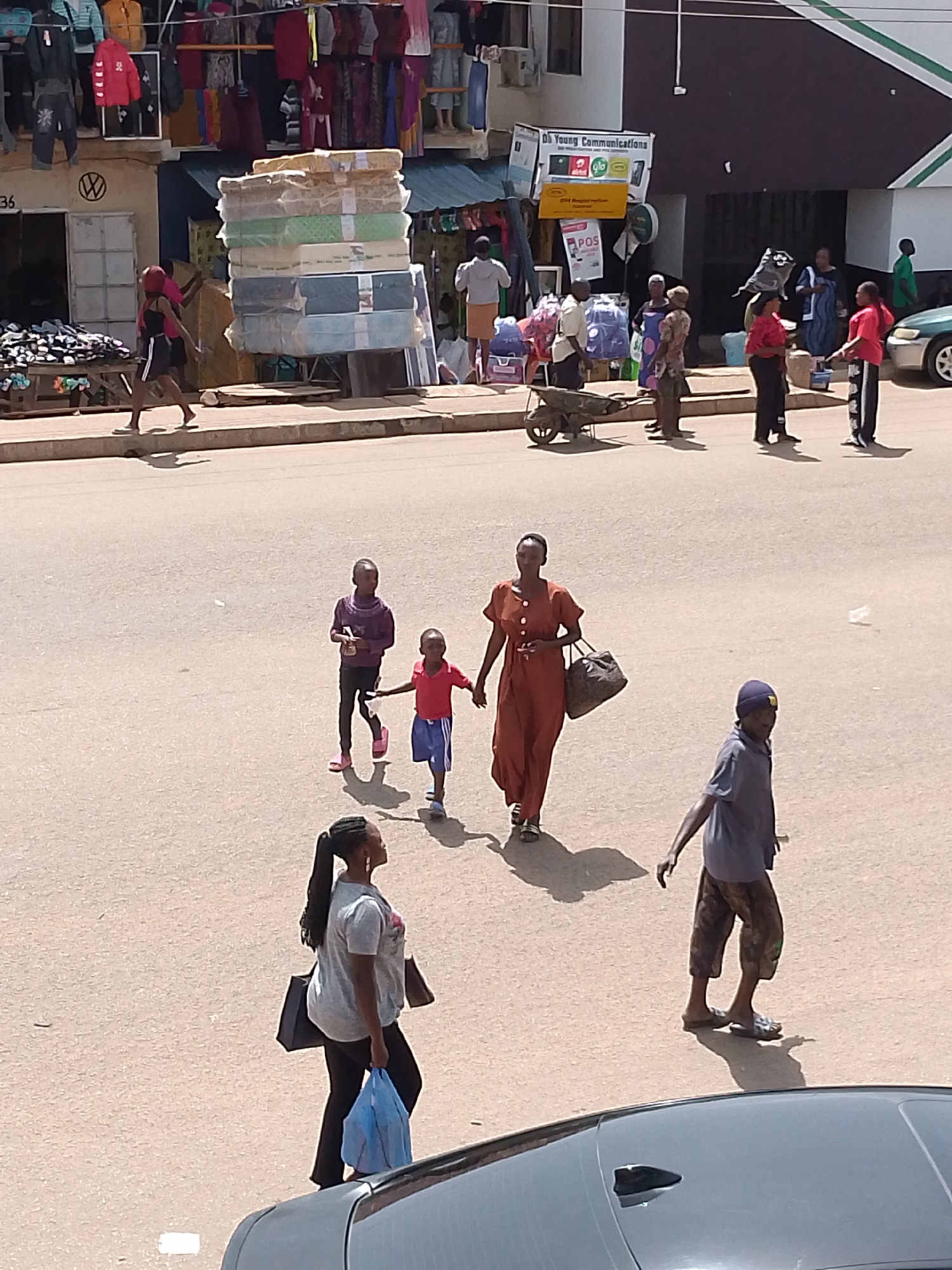This image depicts a busy street scene in Bukuru, Nigeria, likely during daylight hours. The foreground features the roof of a dark-colored car. In the midground, several people are walking and interacting. A woman in an orange dress walks with a brown bag, while a man in a blue shirt and patterned pants, wearing a blue cap, walks in the opposite direction. Two young children, one in a purple sweater and the other in a red shirt and blue shorts, are walking together. Further back, another woman in a grey t-shirt and black pants carries a blue bag. Behind them, a group of people are gathered near stalls displaying merchandise.
The stalls in the background are laden with goods, including stacks of mattresses wrapped in plastic, clothing displayed on racks, and various other items. A wooden cart with a wheelbarrow is visible near the stalls. A sign above one of the stalls reads "DS Young Communications" and displays logos for "Airtel," "Glo," and "9mobile." The scene suggests a marketplace or commercial area where people are shopping or going about their daily activities. The sunlight casts distinct shadows, indicating a bright, possibly warm day. The overall atmosphere appears lively and active.