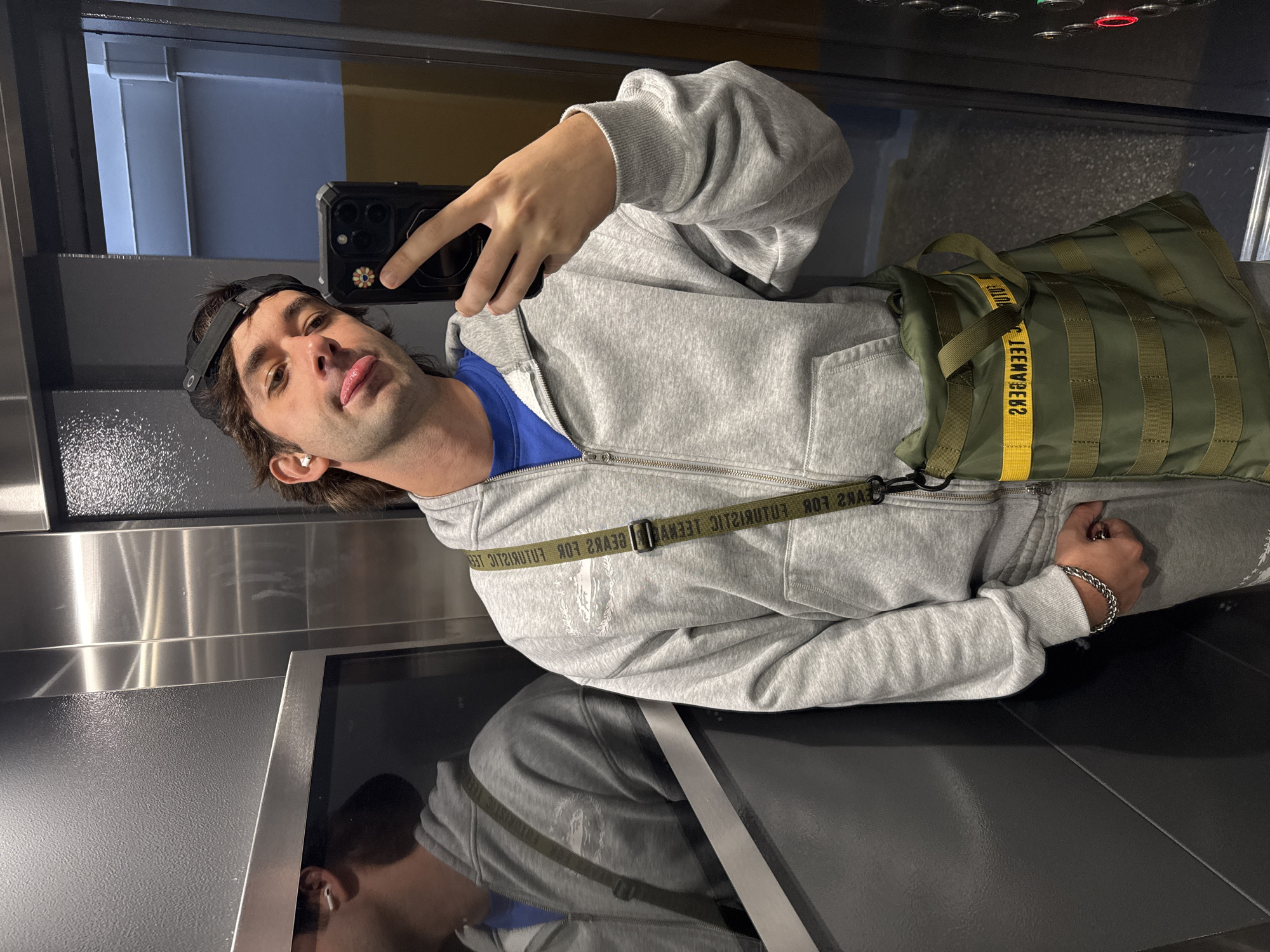 The image shows a young man taking a selfie in what appears to be an elevator. He is wearing a grey zip-up hoodie over a blue t-shirt, grey sweatpants, a black baseball cap, and has wireless earbuds in. He is also carrying a green crossbody bag. The elevator has metallic walls and a mirrored panel reflecting a portion of his body and clothing. The man is sticking his tongue out slightly and holding his phone in his right hand, with his left hand in his pocket. There are elevator buttons visible on the right side of the image, one of which is illuminated red. The scene takes place inside an elevator, not in a specific outdoor city location.