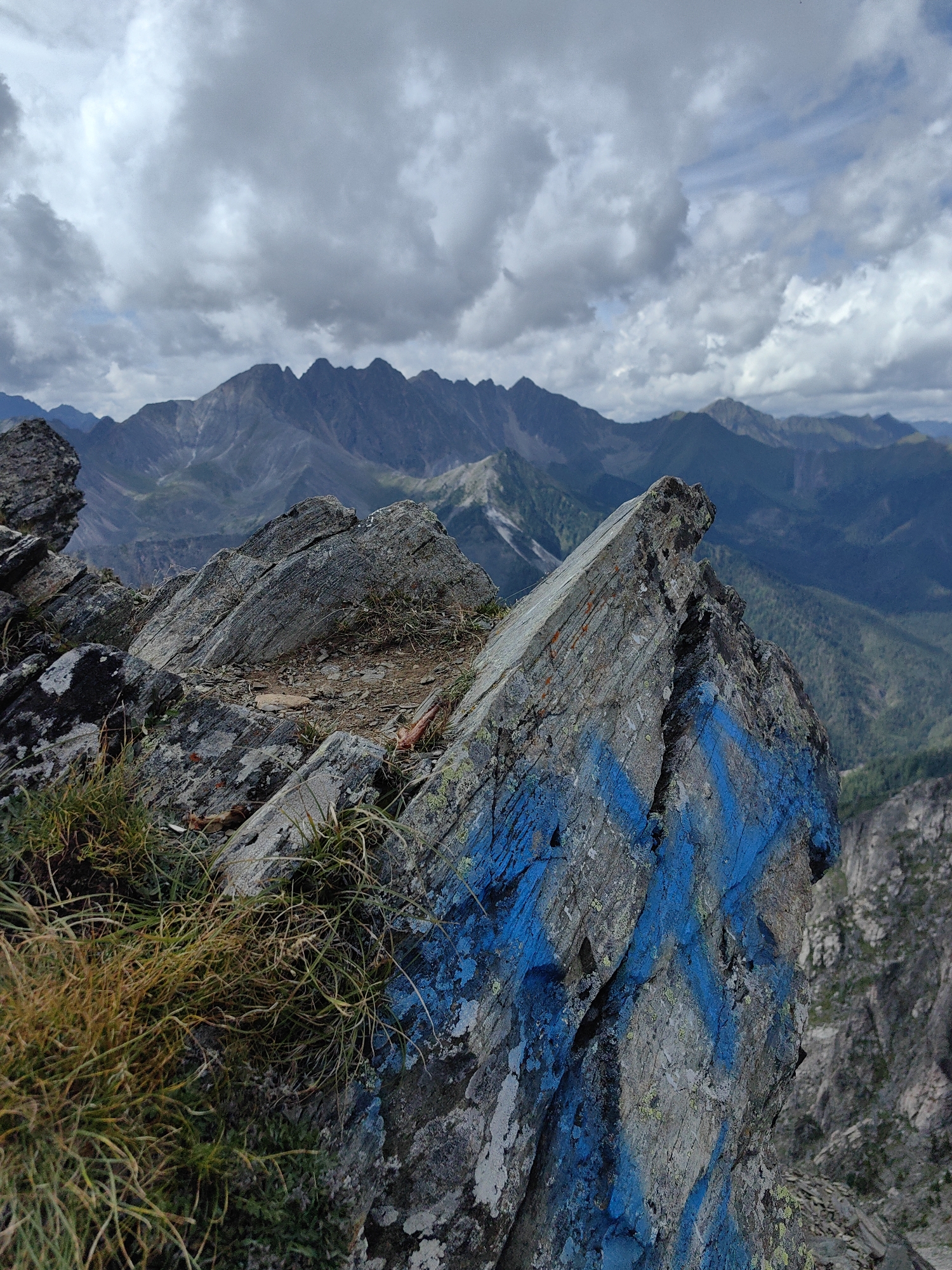 A close-up shot focuses on a jagged, grey rock formation in the foreground, prominently marked with bright blue spray paint markings that form a large "X" and other abstract shapes. Sparse, dry, yellowish-green grass grows from crevices in the rock and the ground around it. In the background, a vast mountain range stretches across the horizon, with rugged peaks and slopes covered in dark green vegetation. The sky above is filled with dramatic, grey, and white clouds, suggesting a potentially overcast or stormy day. The scene appears to be located in a natural, mountainous environment, possibly a hiking trail or a scenic overlook, with the blue markings indicating human presence or a navigational marker. The image is captured from a high vantage point, offering a panoramic view of the surrounding landscape. The overall impression is one of rugged natural beauty juxtaposed with human intervention. The specific location in Arshan, Russia, is not visually evident from the image itself.