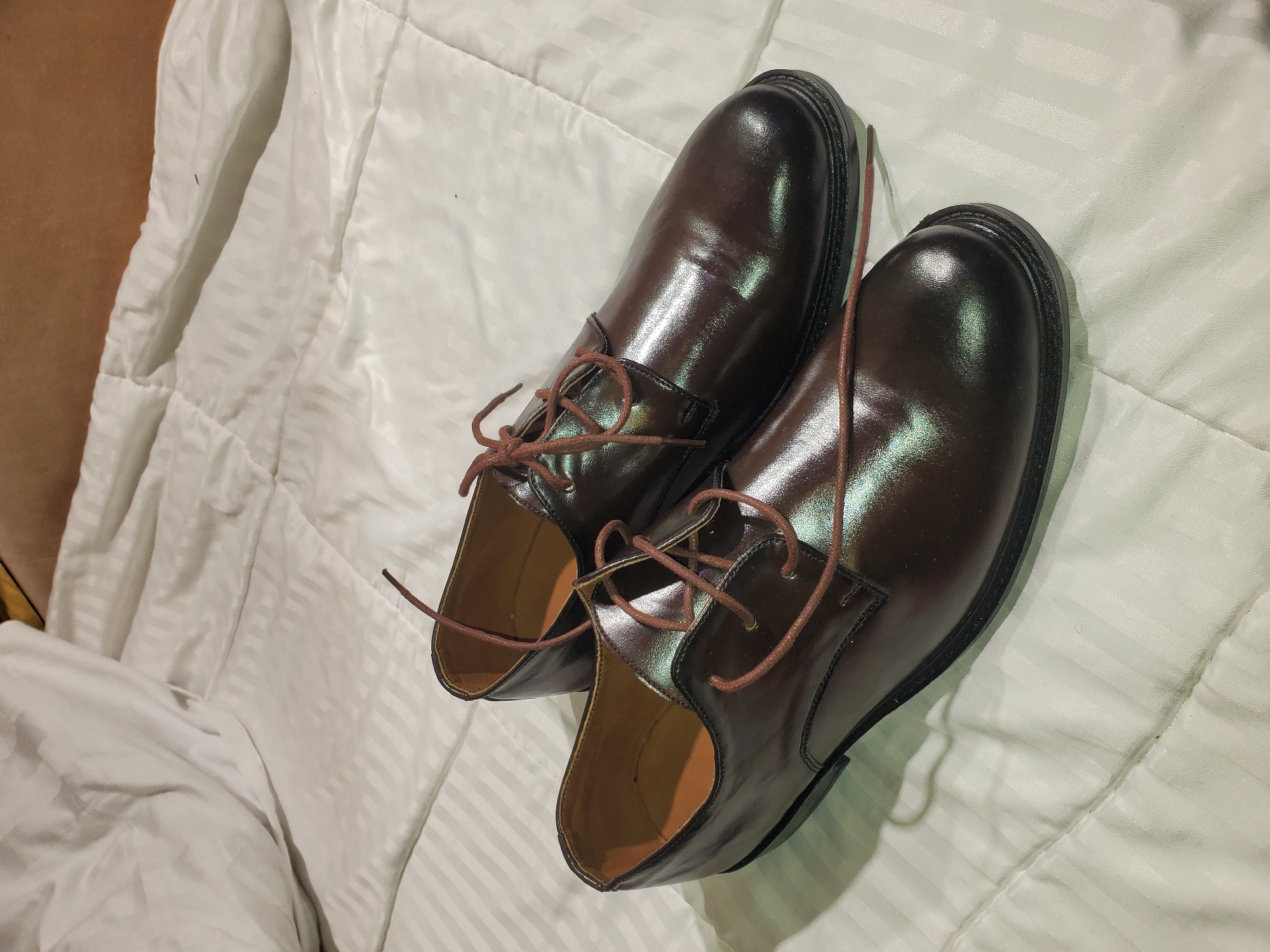 A pair of brown leather dress shoes with brown laces are placed on a white striped bedspread. The shoes are slightly overlapping. The scene is indoors. The location is Unknown, Unknown.
