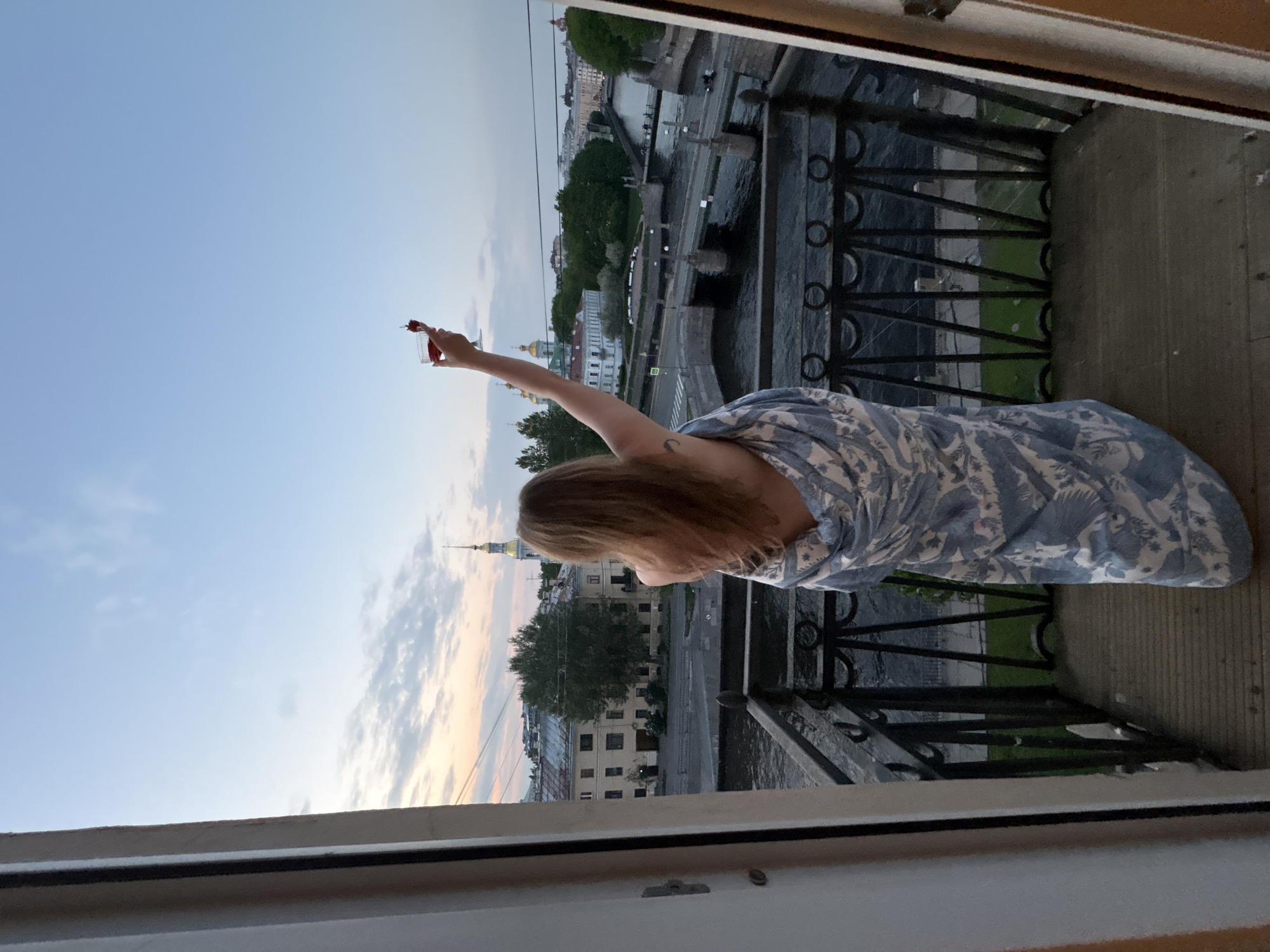 From a high vantage point, likely a balcony or window, a view of St. Petersburg, Russia unfolds. A woman stands, draped in a blue-patterned cloth. She raises her right arm, holding a small glass with a red object in it. The cityscape is visible in the background, including waterways, bridges, buildings, and greenery. A tall, slender spire characteristic of St. Petersburg architecture is also seen. A black iron railing is in the foreground, providing an edge to the balcony.