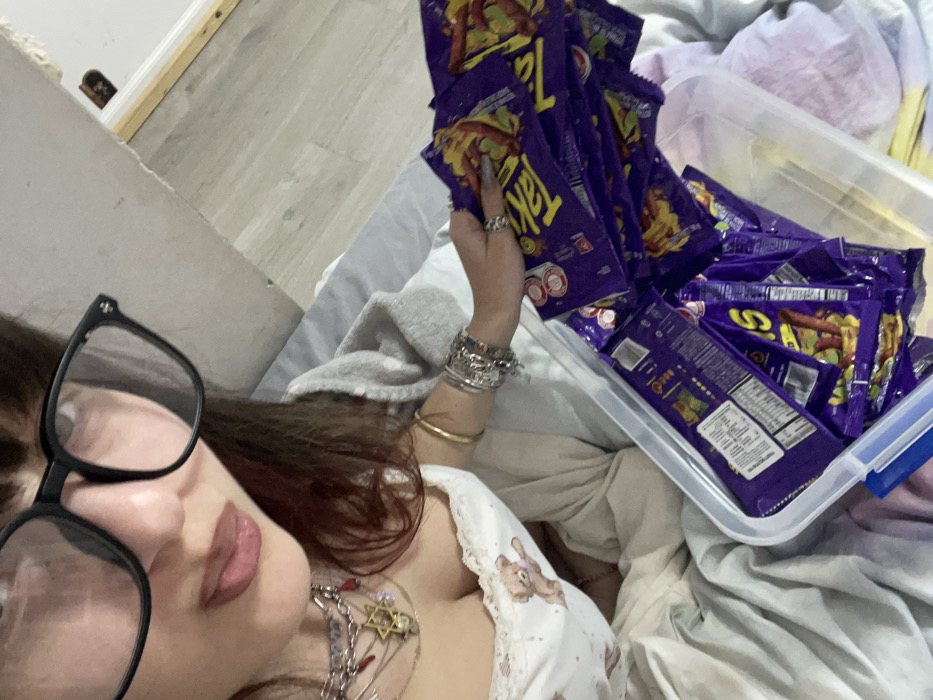 A person wearing large black-rimmed glasses is visible in a selfie. They have multiple bracelets and rings, and several necklaces, including one with a Star of David. They're holding several bags of Takis chips, and a plastic container is filled with more bags of the same chips. The person is lying down amidst bedding.