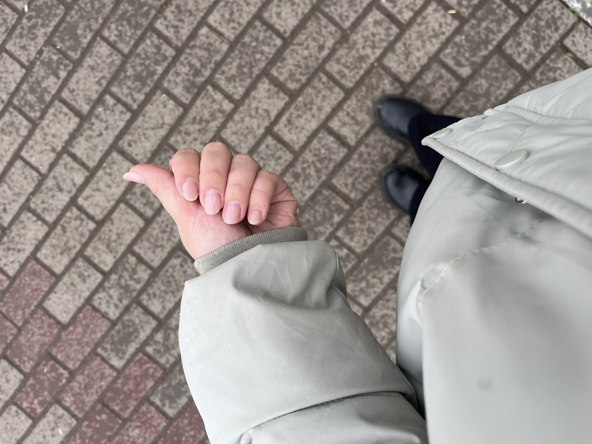 An overhead shot taken at what appears to be an outdoor location in Chaikovsky, Russia. The visible foreground features a person wearing a light-colored puffy jacket and dark shoes, standing on a brick-paved surface. The person's hand is visible, with fingers extended, near the left side of the frame. The ground shows a pattern of rectangular bricks in shades of gray and red. The scene appears to be outdoors, possibly on a sidewalk or walkway. The jacket looks to be buttoned up to their chest with three round buttons. The nails of the person appear to be pink.