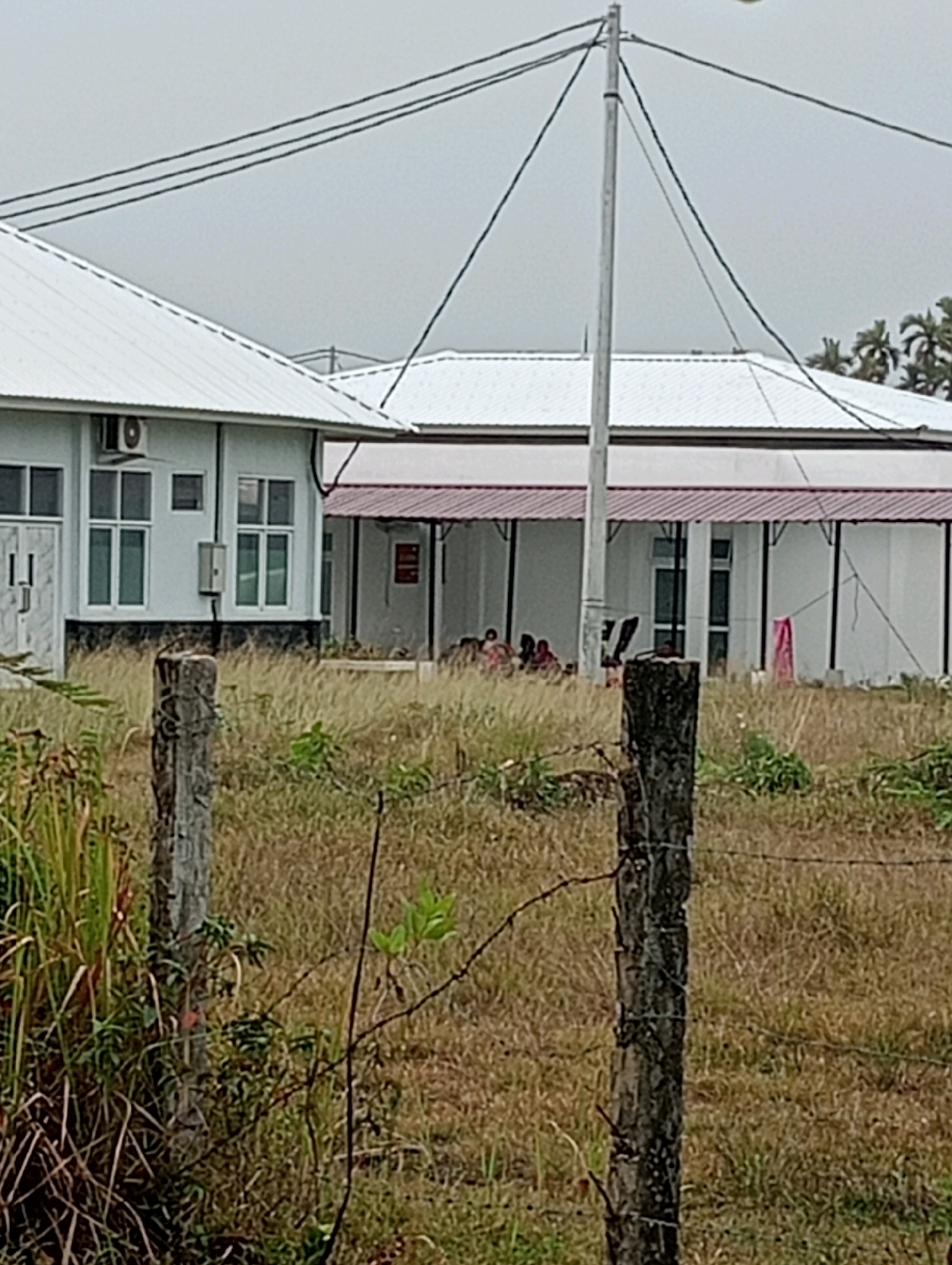 This image depicts the exterior of a white, low-rise building with a white corrugated metal roof, located in Tanjung Pandan, Indonesia. The building features several large windows with white frames, and an air conditioning unit is visible on the left side. A covered walkway with a burgundy roof extends along a portion of the building. In the foreground, a wooden post with barbed wire fencing runs horizontally across the frame, suggesting the property is enclosed. The ground is covered in dry, yellowing grass and some scattered green foliage. A group of people are seated or gathered under the covered walkway of the building. Overhead, power lines crisscross the frame, supported by a tall utility pole. The sky is overcast and gray, indicating a cloudy day. Palm trees are visible in the background on the right side of the image.