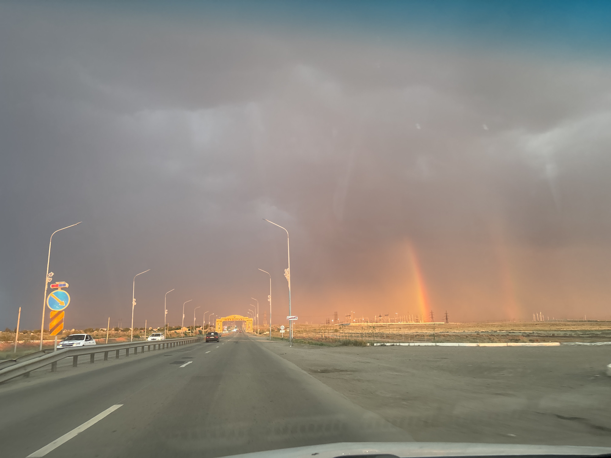 This image depicts a dramatic sky over a highway in Satbayev, Kazakhstan. The sky is dominated by dark, stormy clouds on the left, transitioning to a warm, orange-tinted glow on the right, illuminated by a vibrant double rainbow. The sun is low, suggesting either dawn or dusk, and the light creates a strong contrast between the dark clouds and the illuminated horizon.
The scene is a highway, viewed from the perspective of a vehicle. Cars are visible on the road, moving away from the viewer. On the left side of the highway, there is a railing and a traffic sign with a blue circular symbol and a yellow arrow pointing down and to the right, indicating a specific lane change or turn. Streetlights line the highway, casting a warm glow.
In the distance, a large, archway-like structure is visible, spanning across the road. This structure appears to be an entrance gate or monument, with yellow ornamentation and text. Beyond the gate and stretching towards the horizon, there is a flat, open landscape dotted with industrial elements, including power lines and wind turbines, hinting at the area's infrastructure. The ground along the roadside is a mixture of dirt and gravel.
The overall mood is one of awe and perhaps a sense of impending weather change, given the juxtaposition of the storm clouds and the bright rainbow. The visible text on the archway is "САТБАЕВ" in Cyrillic letters, confirming the location.