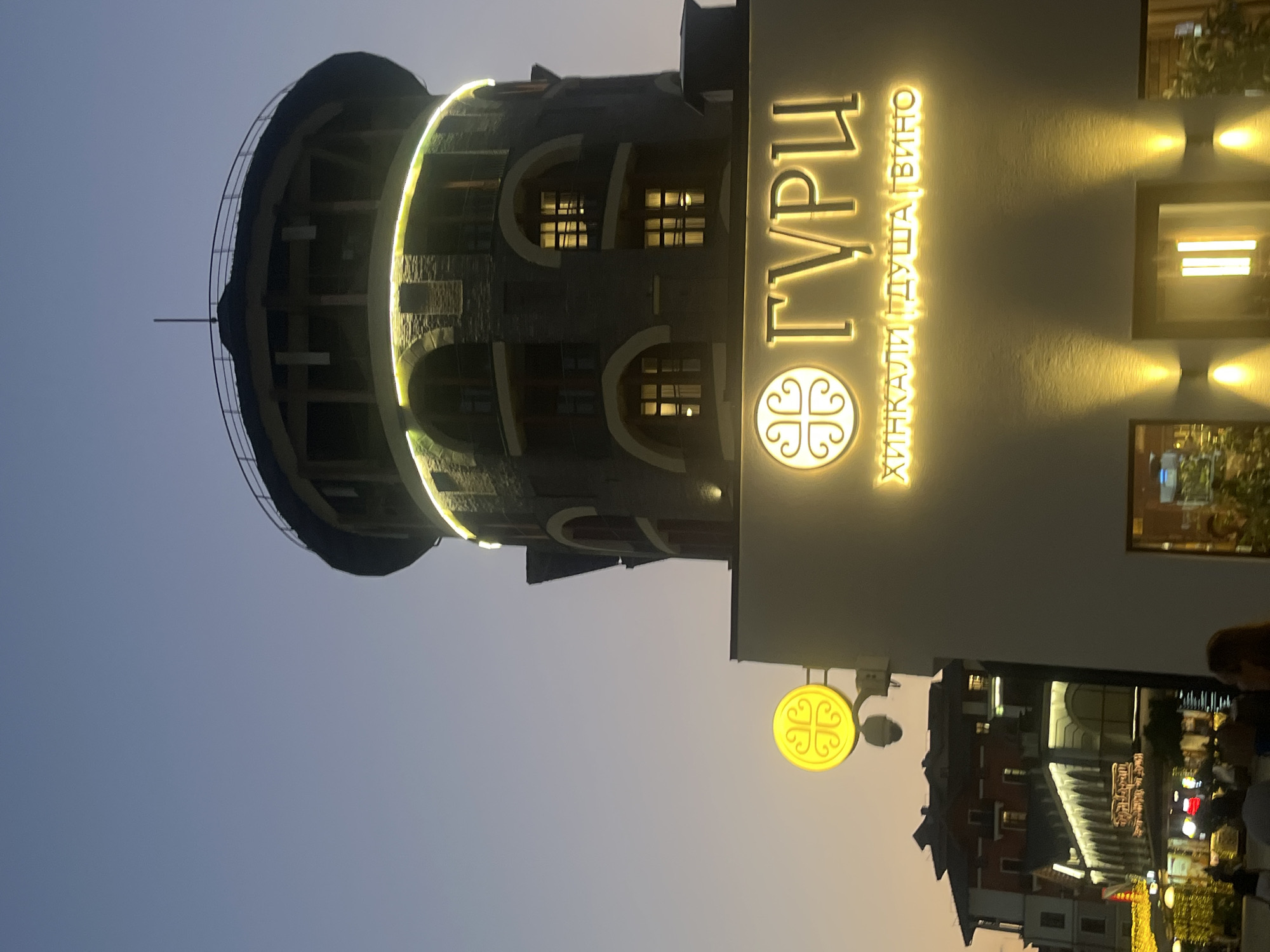 The image captures a nighttime scene in Estosadok, Russia, focusing on a multi-story building with a circular tower. The tower is illuminated with a ring of yellow lights near its top and features stone construction with arched windows. Below the tower, a white building displays a prominent sign with illuminated yellow text. The sign features a circular emblem and the Cyrillic words "ГУРЦ" above smaller Cyrillic text that reads "ХИНКАЛИ ДУША ВИНО." To the left of the main building, a separate circular yellow sign with a decorative motif is mounted on a pole. In the background, other buildings with illuminated facades and rooftops are visible, suggesting a lively commercial or entertainment district. The sky above is a gradient of deep blue to orange, indicating dusk or twilight. Several indistinct figures are present in the foreground and midground, suggesting pedestrian activity.