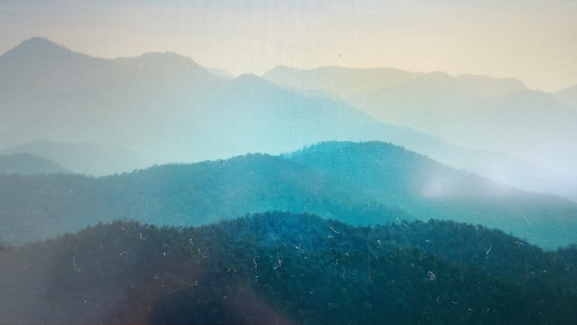 The image depicts a panoramic view of layered, misty mountains. The foreground mountains are dark green and densely covered with trees, indicating a forest. Behind these, several subsequent layers of mountains recede into the distance, becoming progressively lighter and more obscured by fog or haze, transitioning through shades of teal, light blue, and pale yellow towards the sky. The sky is a soft, gradient of light yellow and pale blue, suggesting either sunrise or sunset. There are no people or visible man-made structures in the image. The scene evokes a sense of depth and atmospheric perspective. This landscape is consistent with the mountain ranges found in and around Smolyan, Bulgaria, known for its mountainous terrain and frequent fog.