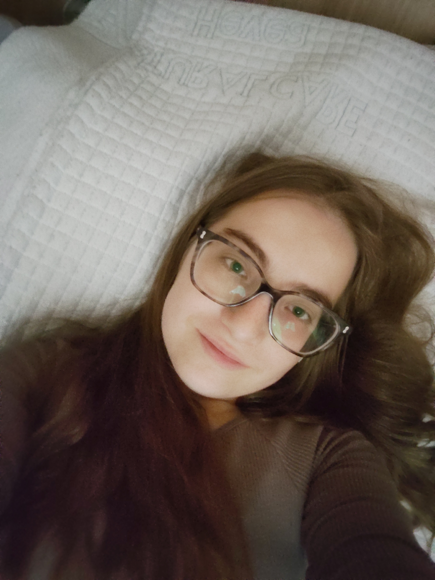 A young woman with light skin, brown hair, and glasses is lying on a white, quilted surface, possibly a bed or large pillow. She is wearing a brown long-sleeved shirt. The quilted surface has a repeating pattern of squares and indistinct embossed lettering. The image is a selfie taken from above. The location is not discernible from the visual information provided. There is no evidence to indicate a location in the city of Sofia, Bulgaria.