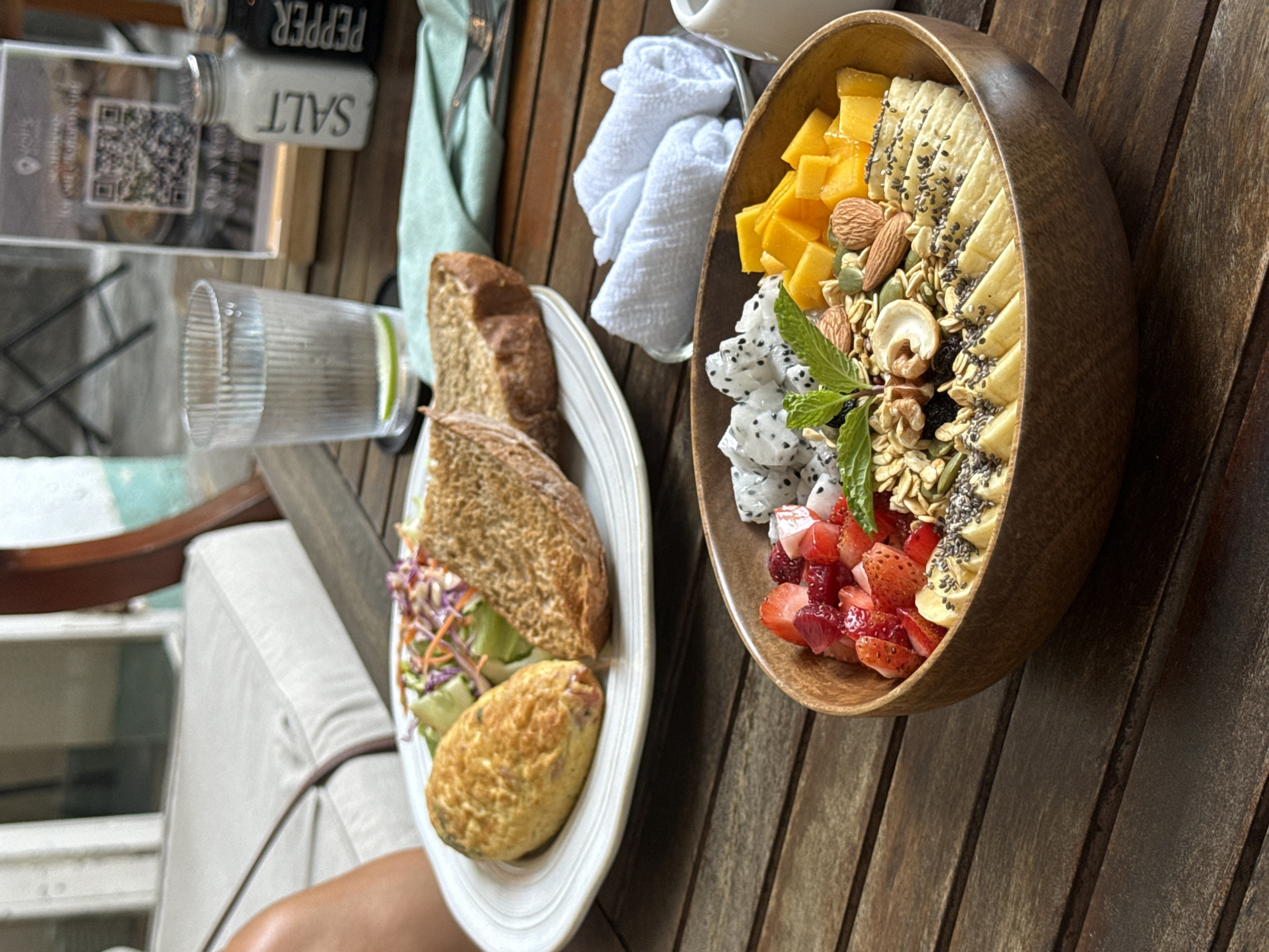 On a wooden table outdoors in Đà Nẵng, Vietnam, a white plate holds a halved sandwich, mixed salad, and oval-shaped food item. Next to it, a wooden bowl is filled with cut mango, strawberries, dragon fruit, banana slices sprinkled with seeds, granola, and nuts, garnished with a sprig of mint. Nearby, a salt shaker, a glass with liquid, and a rolled-up white towel are present. A chair with a white cushion is visible in the background.