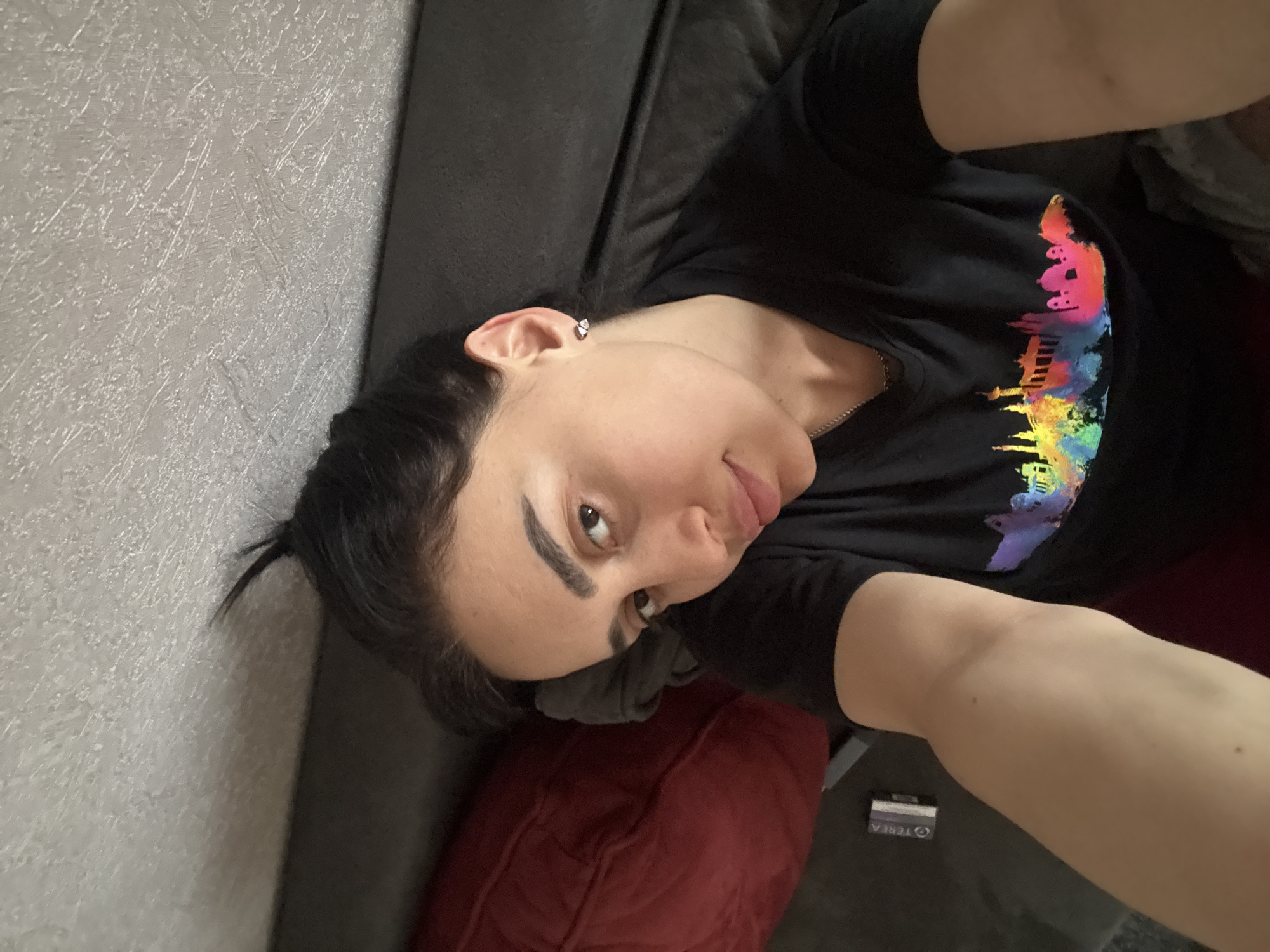A woman is reclining on a gray sofa. A dark gray sofa is positioned along a wall with textured light gray wallpaper. A red pillow is under her head. The woman has dark hair pulled back and is wearing an earring in her left ear. She is wearing a black T-shirt with a colorful silhouette of buildings on it. Her left arm is partially visible and holding a camera phone. A pack of TEREA cigarettes is visible near her left arm. The location in Tbilisi, Georgia is not discernible from this image.