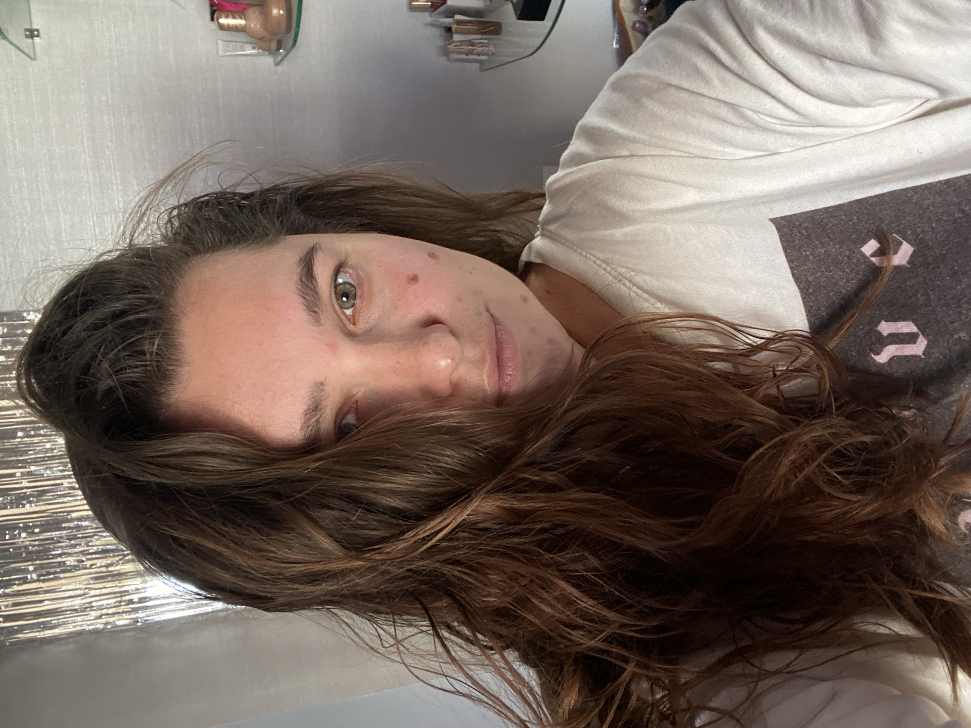 A woman with brown, wavy hair is taking a selfie. She is wearing a white t-shirt with a graphic design. Her hair is covering the left side of her face. She has green eyes and some blemishes on her skin. In the background, there is a silver, sparkly curtain, a shelf with bottles of makeup and a white wall. The location is not specified to be in the city of Perm, Russia.