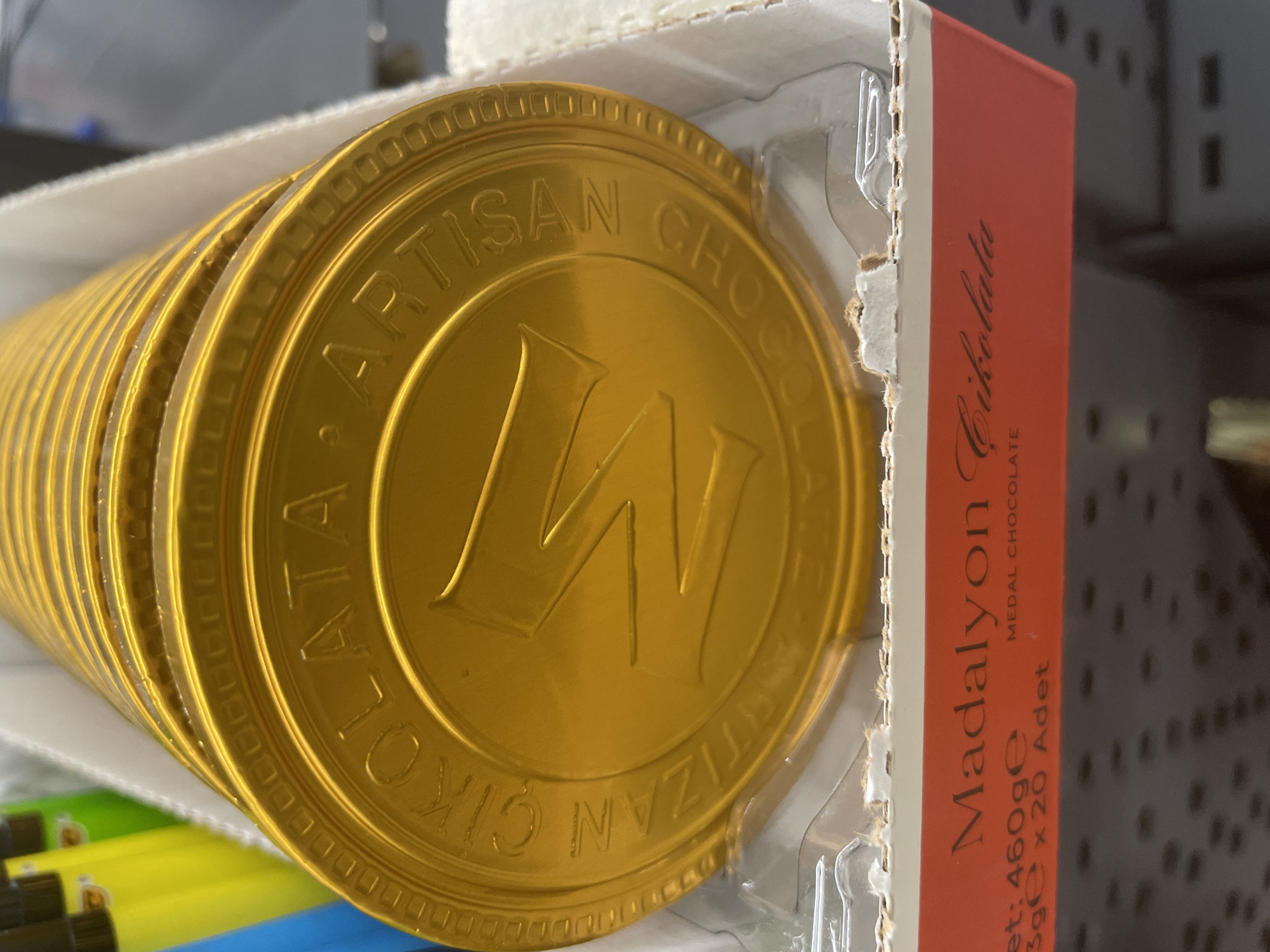 The image shows a close-up, slightly angled view of stacked gold-colored chocolate medals, likely a product packaging. The medals are circular with embossed text and a large, stylized "M" in the center. The text around the edge of the visible medals appears to read "ÇIKOLATA ARTISAN CHOCOLATE". Below the chocolate medals, a red cardboard band with white text is visible. The text on this band reads "Madalyon Çikolata" and below that, "et: 460ge", "3ge x 20 Adet", and "MEDAL CHOCOLATE". The packaging is in a white cardboard box. In the background, blurred yellow and green objects are visible, possibly other products or packaging materials. The lighting suggests an indoor environment, likely a retail setting. There is no indication of the city of Yunus Emre, Turkey within the image itself.