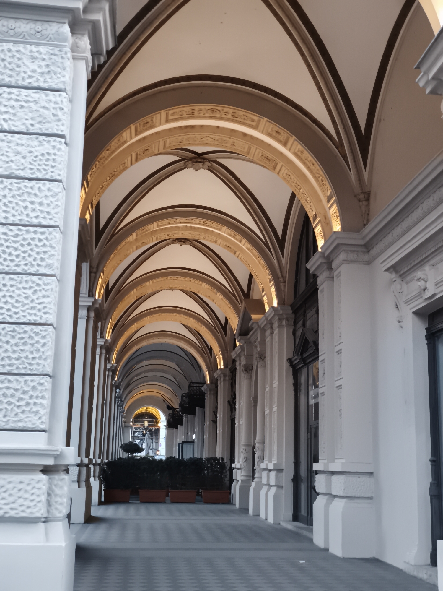The image shows a perspective view of a covered walkway or arcade with a series of arched ceilings. The architecture is ornate, with decorative moldings along the arches and columns. The walkway recedes into the distance, leading to an arched opening at the end of the street. On the right side of the walkway, there are windows and doors, suggesting commercial establishments. On the left side, the wall has a textured finish and is punctuated by pilasters. The ground is paved with a checkered pattern. In the foreground, there are planters with greenery. In the distance, there is scaffolding visible, indicating ongoing construction or renovation. The lighting suggests it is either early morning or late evening, with artificial lights illuminating the arches. This scene is located in the Graben in Vienna, Austria, a famous shopping street known for its arcades and historic architecture. There are no people visible in the image.