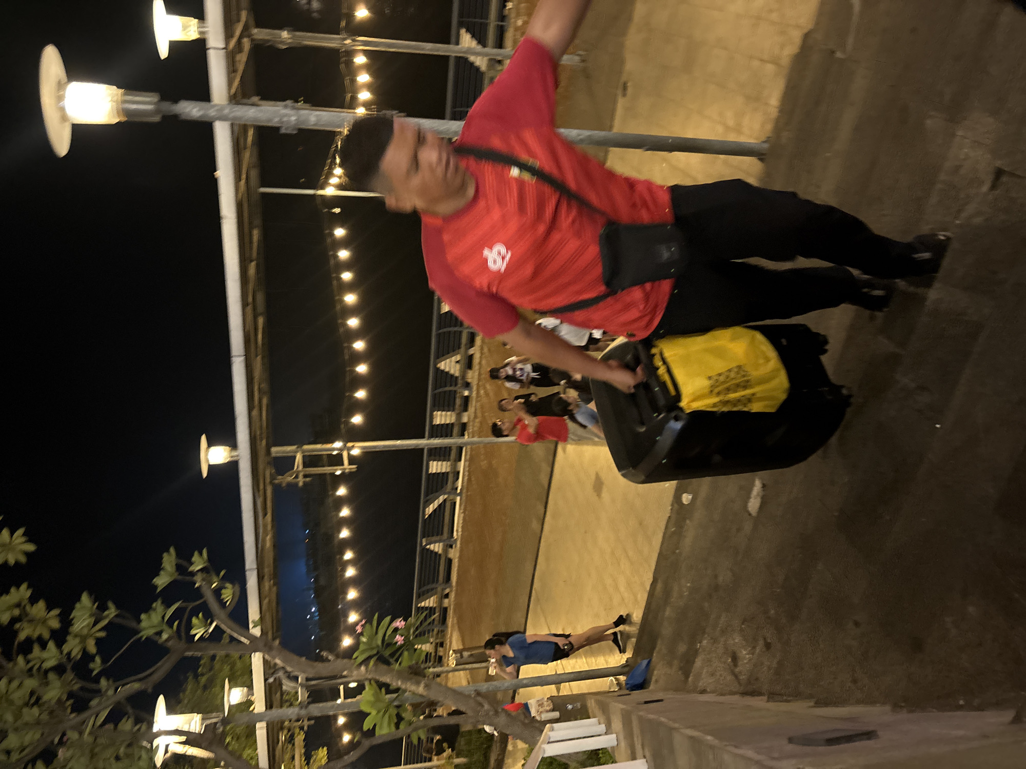 At night, on an outdoor staircase in Ancol, North Jakarta, Indonesia, a man in a red t-shirt and black pants is walking up the stairs. He is carrying a large black speaker with a yellow bag attached to it. There are other people in the background, and string lights are visible along a railing. The scene is lit by streetlights and the string lights.