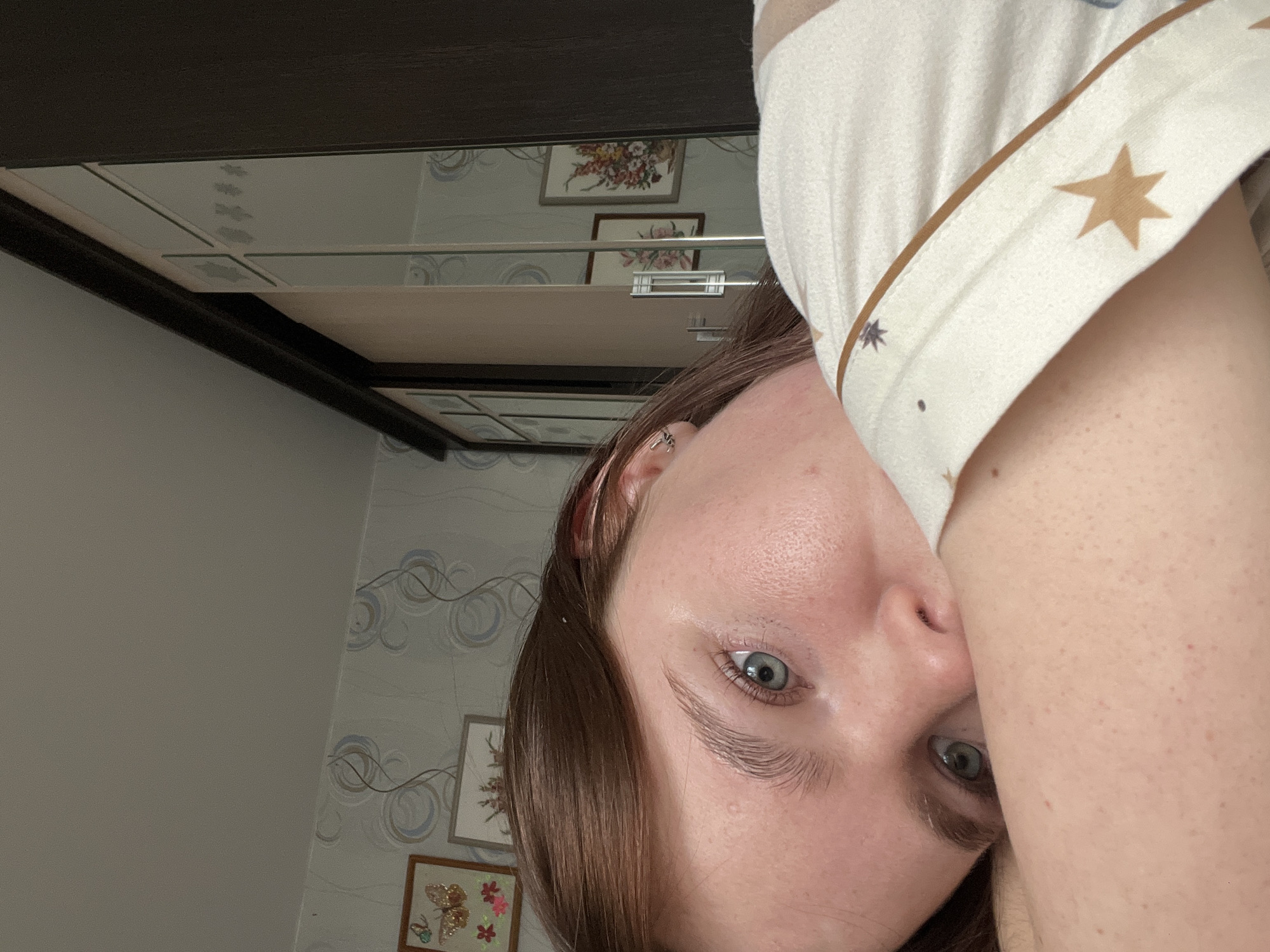 A close-up, tilted shot of a person's face is visible, with light brown hair and blue eyes. The person is wearing a light-colored garment with brown star patterns. The background shows a section of a room. Wallpapers with circular patterns are present along with framed artwork hanging on the wall. A dark-colored piece of furniture, possibly a cabinet, is positioned above. There are no discernible details to confirm its location within Chaikovsky, Russia.