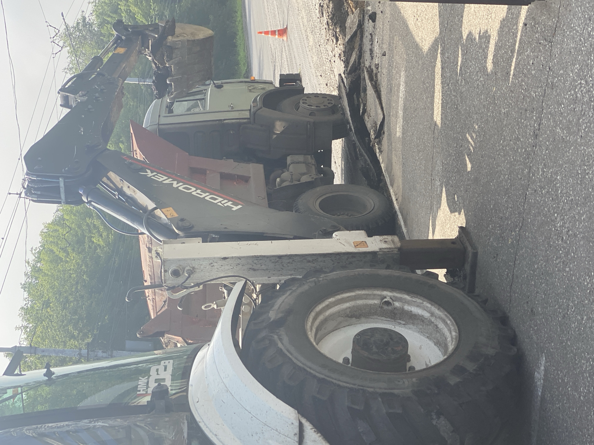 The image depicts a construction scene in Novokuznetsk, Russia, focusing on heavy machinery and road repair. In the foreground, the large tire and white fender of a backhoe loader are visible on the left. The loader's arm, with a bucket filled with dirt, is extended towards a dump truck positioned in the center of the frame. The dump truck is light green with a dark orange bed. The word "HIDROMEK" is prominently displayed on the side of the loader's arm. The road surface shows signs of excavation, with broken asphalt and piles of dirt. A bright orange traffic cone is visible on the right side of the road, suggesting ongoing work or a closed area. In the background, lush green trees line the area beyond the road, and utility poles with overhead wires are present. The sky is overcast. There are no people visible in the image.
