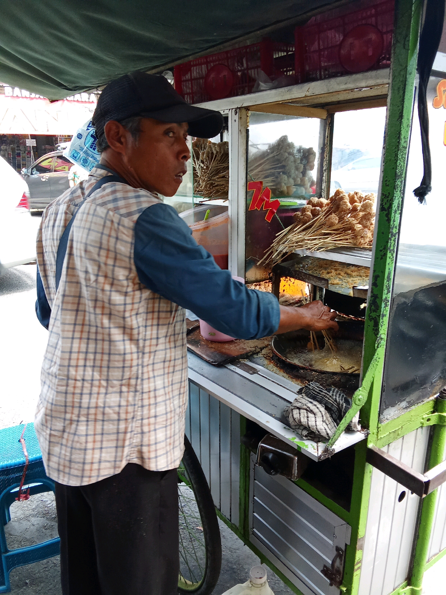 An Indonesian man wearing a plaid shirt and a black cap is standing at a food cart in Banjarnegara, Indonesia. He is in the process of frying food, with his hand submerged in a pan of oil. The food cart is filled with skewers of fried food. The background shows a street scene with a silver car. The cart is painted green and white, with a metal frame. There is a blue plastic chair to the left of the cart, and a bicycle wheel is visible to the right. The scene appears to be outdoors, possibly on a street or market.