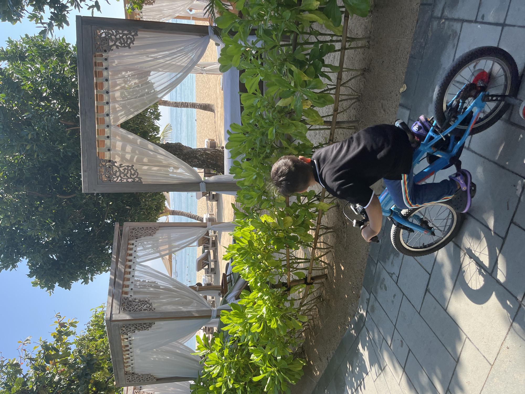 A young boy rides a blue bicycle on a paved pathway beside a tropical beach resort in Kuta Selatan, Indonesia. The boy is wearing a black shirt and blue jeans with rainbow stripes. He is facing away from the camera, looking towards the beach. The pathway is paved with large, light-colored rectangular tiles, and there is a shadow cast by the boy and his bicycle. To the right of the pathway is a manicured hedge of bright green leafy plants, supported by a bamboo fence. Beyond the hedge is a sandy beach with white cabanas featuring white draped curtains and ornate wooden detailing. The ocean is visible in the background, with a clear blue sky above. There are palm trees and other lush foliage visible throughout the scene, characteristic of a tropical location.
