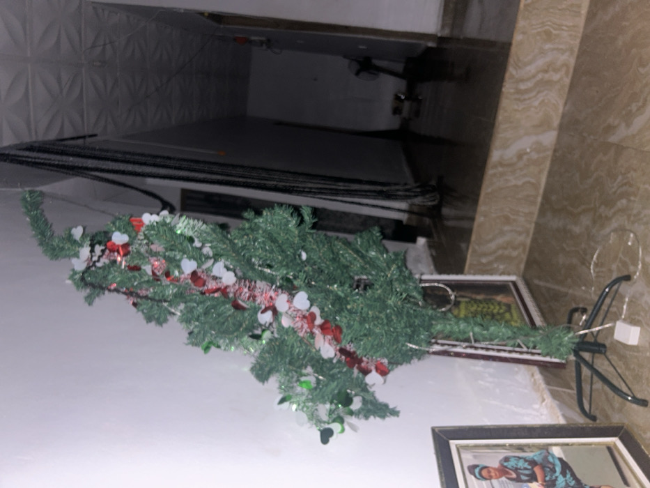 A fallen artificial Christmas tree with heart-shaped decorations lies on its side. The tree's metal stand is visible, along with framed pictures nearby. Above, electrical cords run across a wall with patterned panels. The background shows a hallway with dark and light tiles. The perspective makes the scene appear tilted.