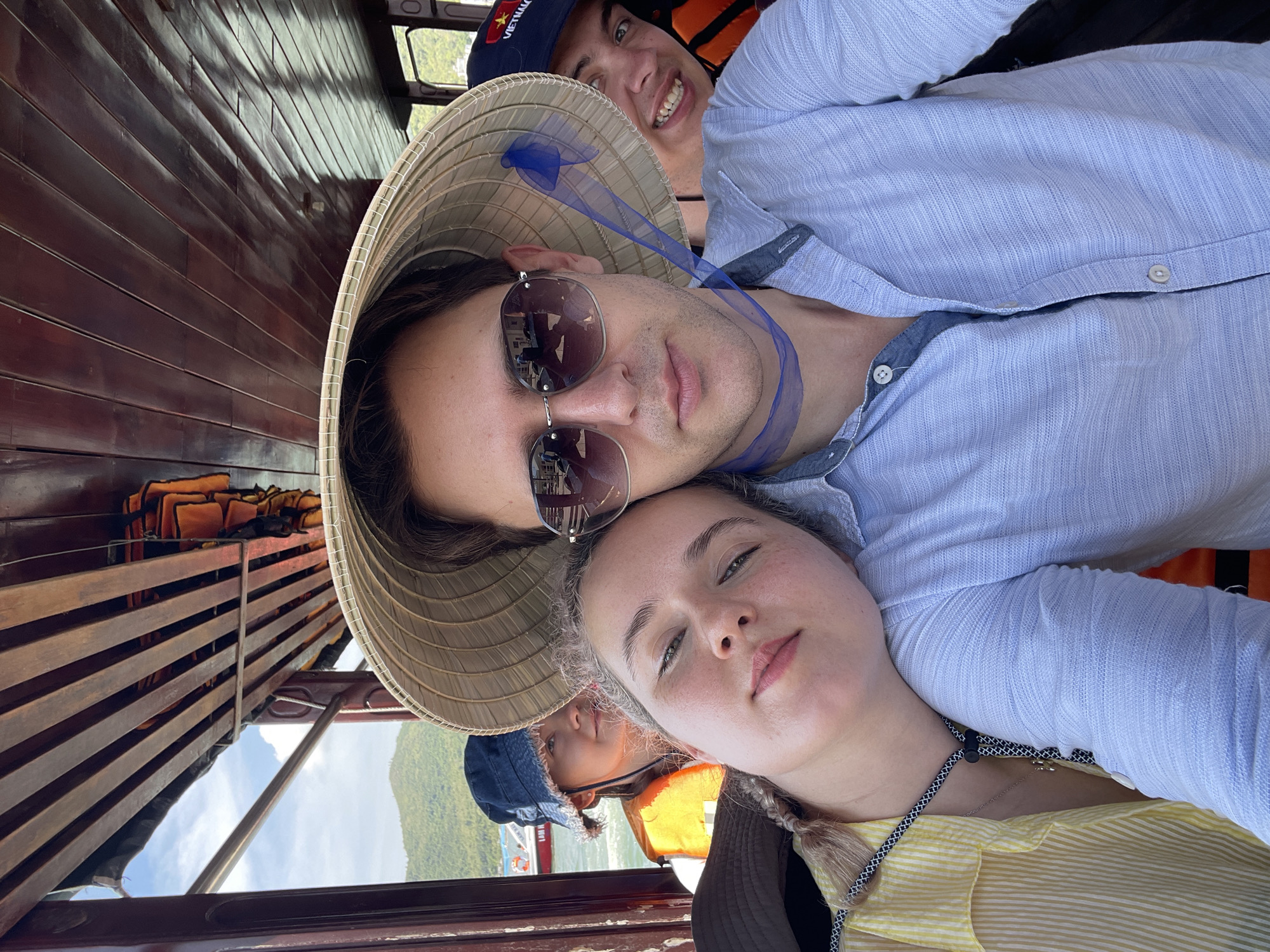 Three people are in a boat, taking a selfie. A woman in a yellow striped shirt is in the foreground and has her head under the chin of a man wearing sunglasses and a conical hat. Another man is behind them, wearing a Vietnam hat and a life vest. The boat is in the city of Nam Nha Trang Ward, Vietnam. Life vests are stored on a shelf above them.