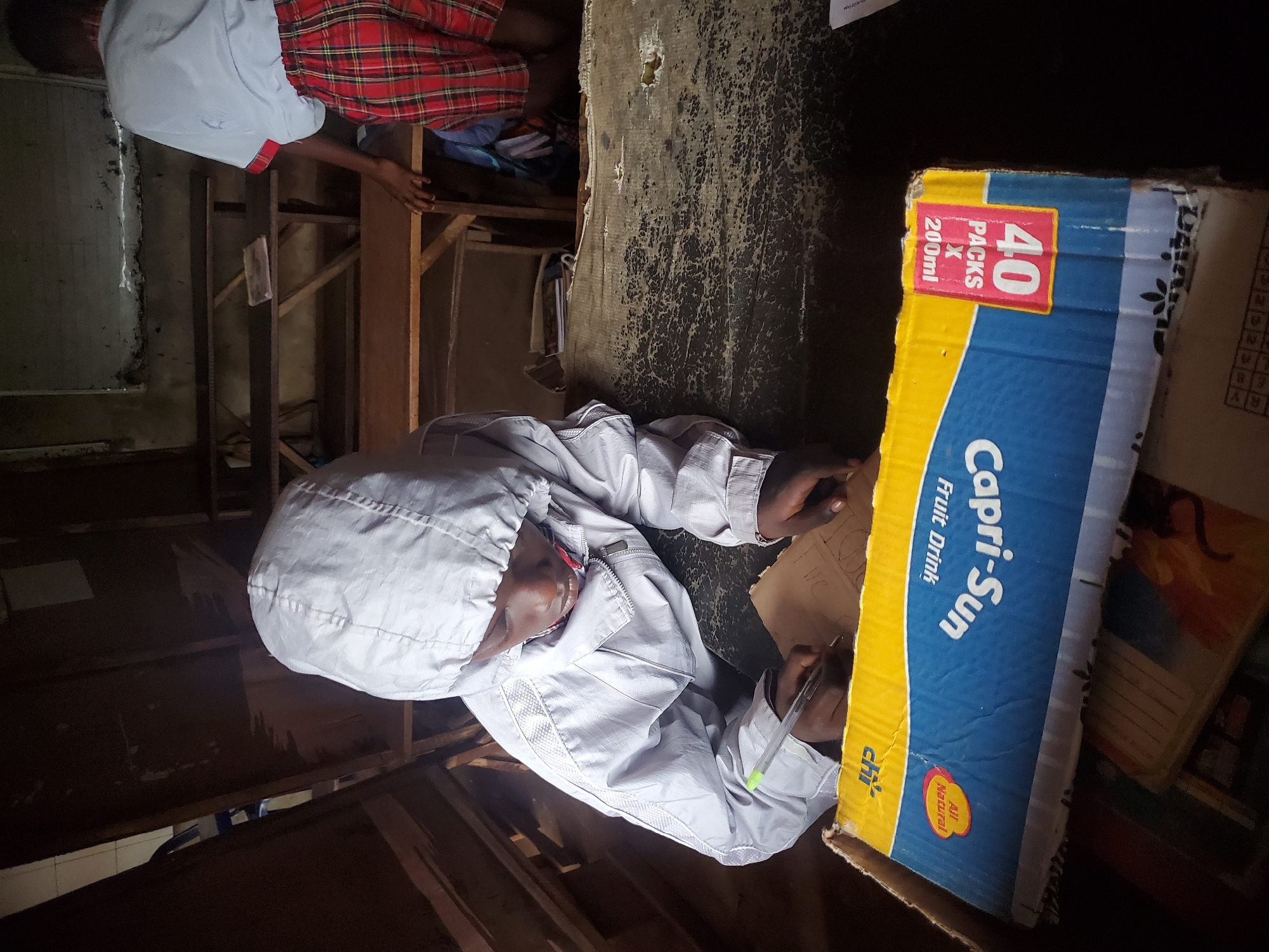 In Ukwu-Abwa, Nigeria, a person wearing a light-colored hooded jacket is writing on a piece of paper. The person is using a pen and appears to be focused on the task. The paper is resting on top of a Capri-Sun fruit drink box, which indicates it contained 40 packs of 200ml drinks. The environment looks to be a dimly lit interior. Another person can be seen in the background, wearing a red and white plaid pattern shirt. The space seems confined and possibly makeshift.
