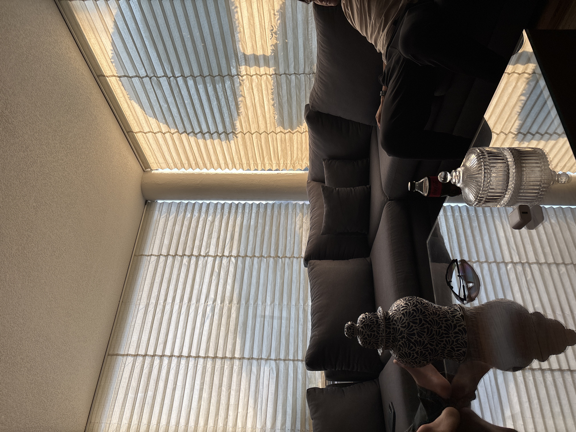 An indoor scene in San Bernardino Tlaxcalancingo, Mexico, shows a modern living area bathed in natural light. The image is oriented sideways, but if rotated, a large window and sloped ceiling on the left are covered with light-colored pleated blinds, creating vertical and horizontal lines. Bright sunlight streams through the blinds, casting distinct shadows of what appears to be foliage onto them, indicating a sunny daytime.
A person is partially visible on the right, seated on a dark grey, plush sofa with multiple cushions. They are wearing a light-colored top and dark pants, with bare feet showing. In front of the sofa is a clear glass coffee table, which strongly reflects the blinds and other room elements.
On the glass table, several objects are arranged: a tall, clear glass decanter with a fluted design and a stopper; a small red-capped bottle, identifiable as "Coca-Cola" from its label; a pair of dark sunglasses; and a decorative urn or vase with an intricate black and white/cream geometric or floral pattern. The wall on the left features a light-colored, textured finish. The overall ambiance is bright and relaxed, with strong visual emphasis on light, shadows, and reflections.