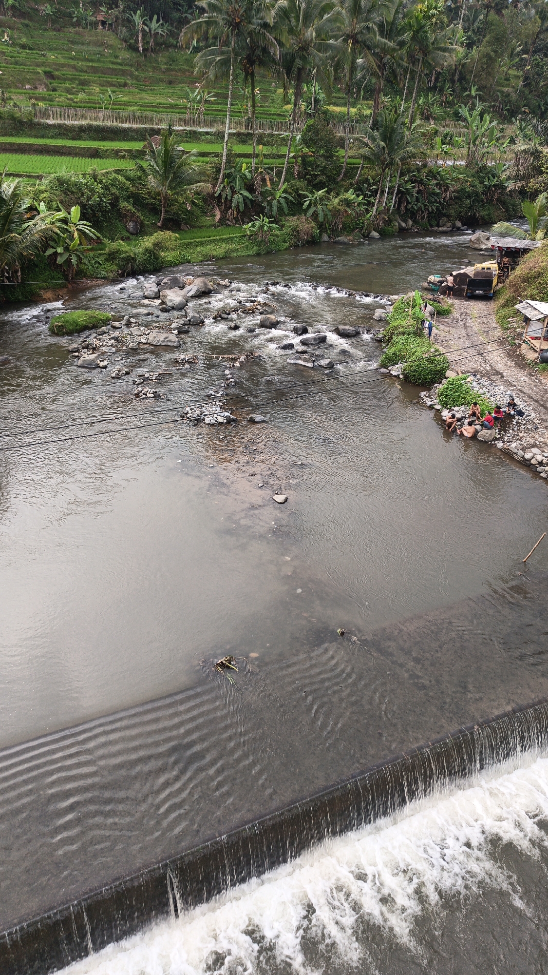 This image captures a scenic view of a river in Cidugaleun, Indonesia, with terraced rice paddies and lush vegetation in the background. A concrete dam creates a small waterfall in the foreground, with foamy white water cascading over the edge. The river flows gently in the middle ground, with scattered rocks breaking the surface. On the right bank of the river, a group of people are gathered, some sitting on the rocky shore and others in the shallow water. A truck and a small shelter are also visible on the bank, suggesting a recreational or work area. The overall impression is one of a tranquil rural landscape, with human activity integrated into the natural environment.