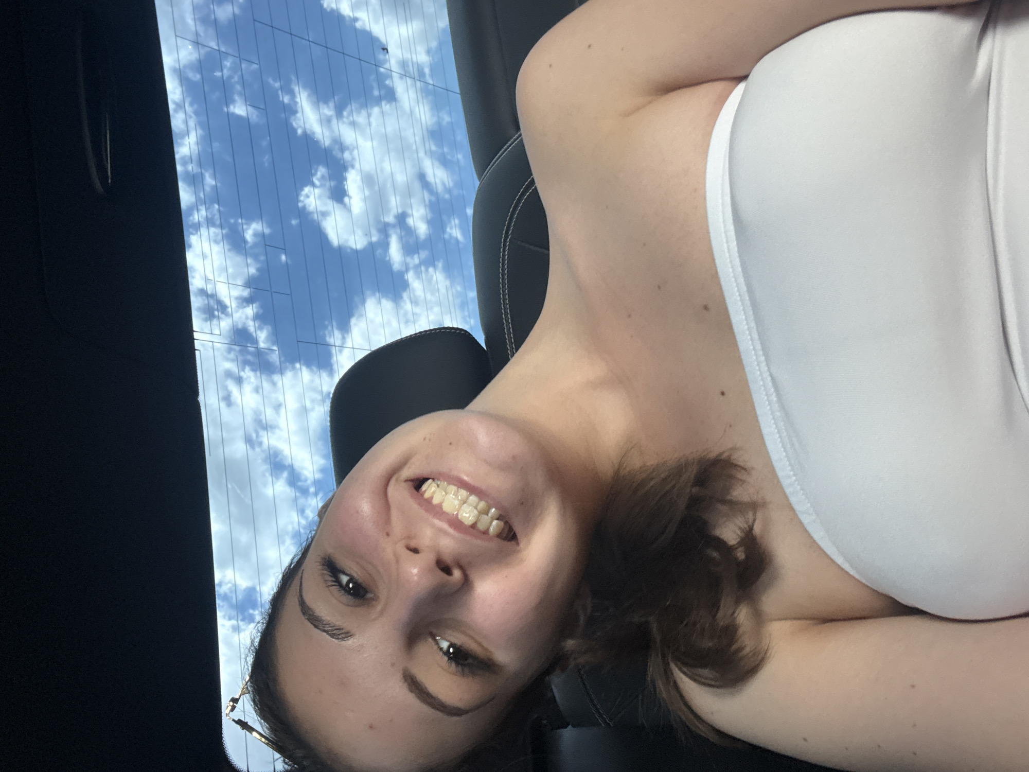 A young woman with dark hair and brown eyes is taking a selfie in a car. She is smiling and wearing a white strapless top. The background shows a blue sky with clouds through the car's rear window. The scene appears to be outdoors, possibly in a city given the presence of a car and what looks like the texture of a car seat. The specific location in the city of Burgas, Bulgaria cannot be determined from the provided image.