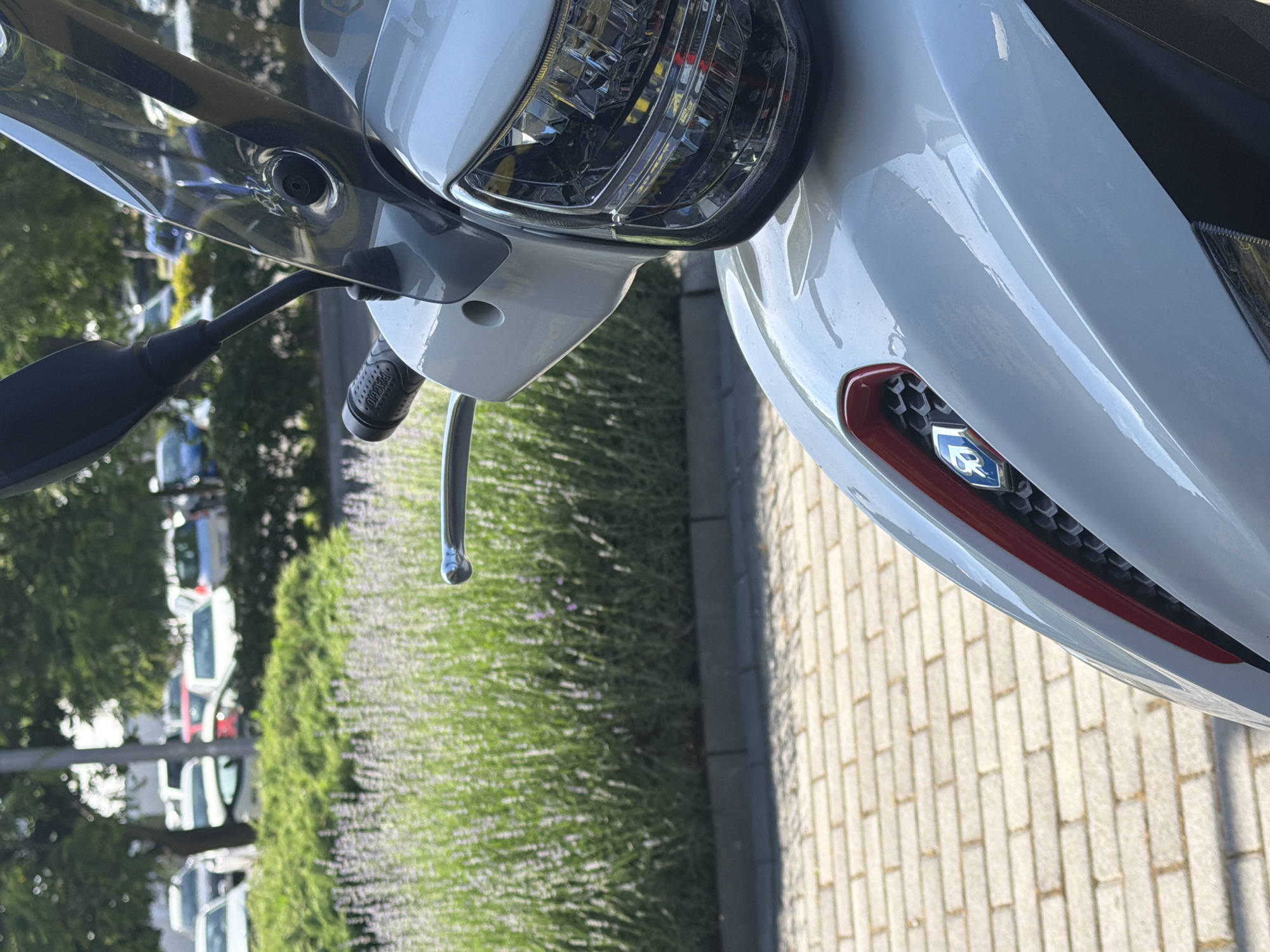 The image shows a close-up of a white scooter parked on a paved surface. In the background, a row of lavender bushes borders a parking lot filled with cars. Trees are visible behind the cars, suggesting an outdoor setting in a city environment. The scooter's headlight and a portion of its bodywork, including a red and chrome accent with a blue logo, are in focus. The scene appears to be a sunny day. The location is identified as Burgas, Bulgaria. There are no people visible in the image, and no actions or interactions are depicted.