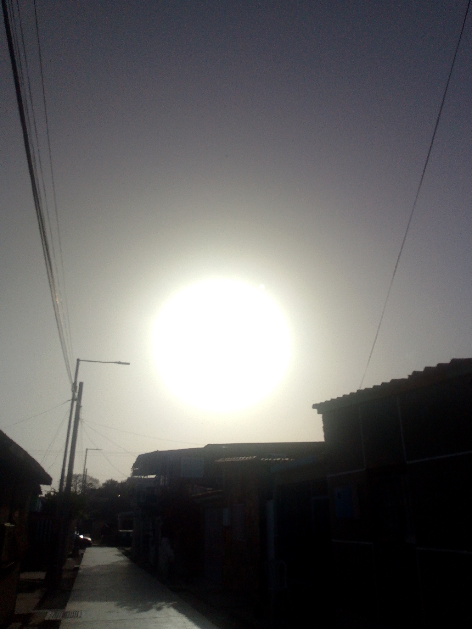 The image captures a sun-drenched street scene in Juan Griego, Venezuela, characterized by extreme backlighting under what appears to be midday or early afternoon conditions. A brilliant, almost blinding, circular light source—the sun—dominates the upper center of the frame, causing significant overexposure and a strong halo effect. The sky around it is a light greyish-white, gradually darkening to a muted grey-blue towards the upper corners, with some visible grain.
The street, possibly a narrow alleyway, is paved and recedes into the distance. Buildings with distinct tiled roofs line both sides, appearing in deep silhouette due to the intense sunlight. These structures are rendered almost entirely black, with architectural details like windows and doors obscured by shadow. On the left side, a tall utility pole supports a streetlamp and numerous overhead wires that crisscross the sky. Additional wires are visible stretching across the upper right portion of the frame.
Towards the end of the visible street, some dark shapes suggestive of trees or bushes are faintly discernible. A small, dark object, possibly a parked car, can be vaguely made out on the left side of the street in the distance. The overall mood is one of stark contrast and intense light. No people or specific activities are depicted, and no visible text is present.