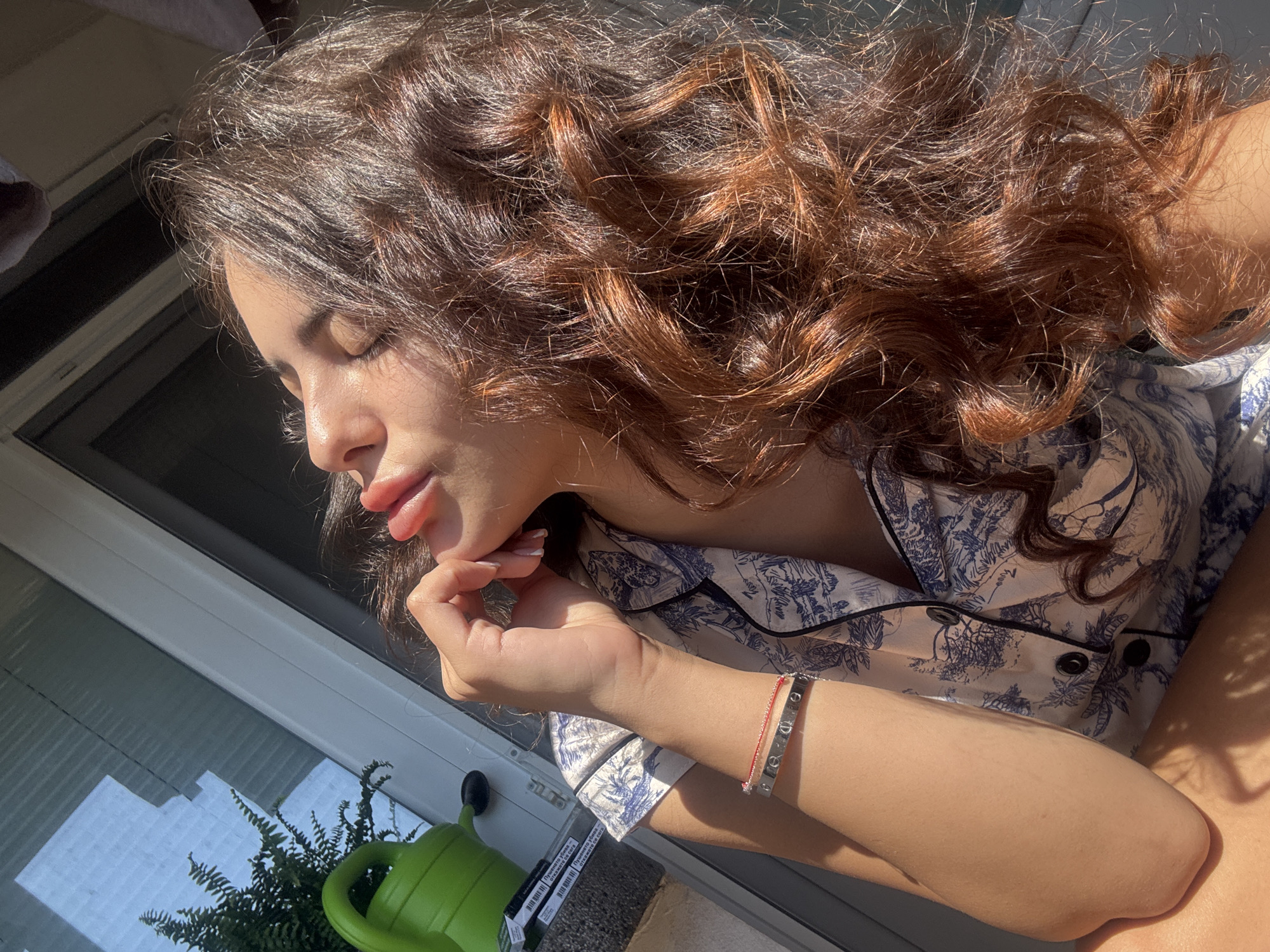 A young woman with long, wavy, brown hair is captured in a close-up, side profile shot, bathed in sunlight. Her eyes are closed, and her head is tilted back slightly, with her lips parted as if basking in the warmth. Her hand is raised, with fingers lightly touching her chin, and she is wearing a bracelet on her left wrist. She is dressed in a pajama top with a blue and white toile pattern. The background suggests an indoor setting near a window, with a green watering can and a plant visible on a surface to the left. The sunlight creates a strong highlight on her face and hair, emphasizing the texture and color of her hair. The scene is not indicative of any specific location within the city of Plovdiv, Bulgaria.