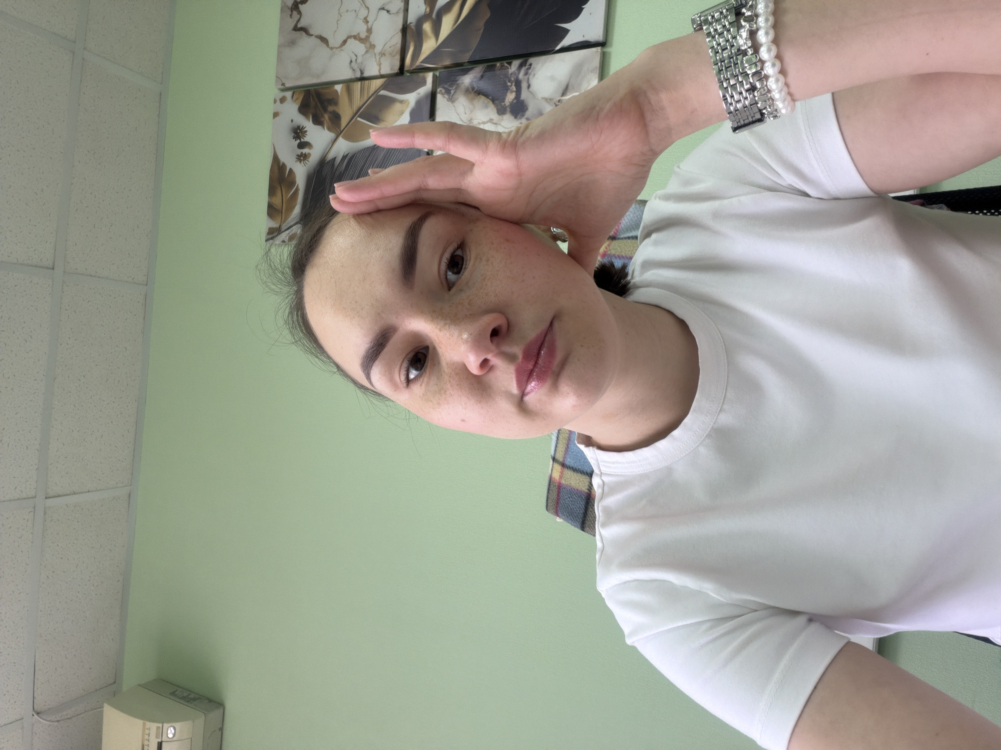 A person with light skin, brown hair, and freckles is posing for a selfie. They are wearing a white t-shirt and a bracelet with silver and pearl elements on their left wrist. Their left hand is raised to their forehead. The background consists of a light green wall, a patterned picture, and a white drop ceiling. The photo was likely taken indoors in Chaikovsky, Russia. There is no apparent indication of the exact location.