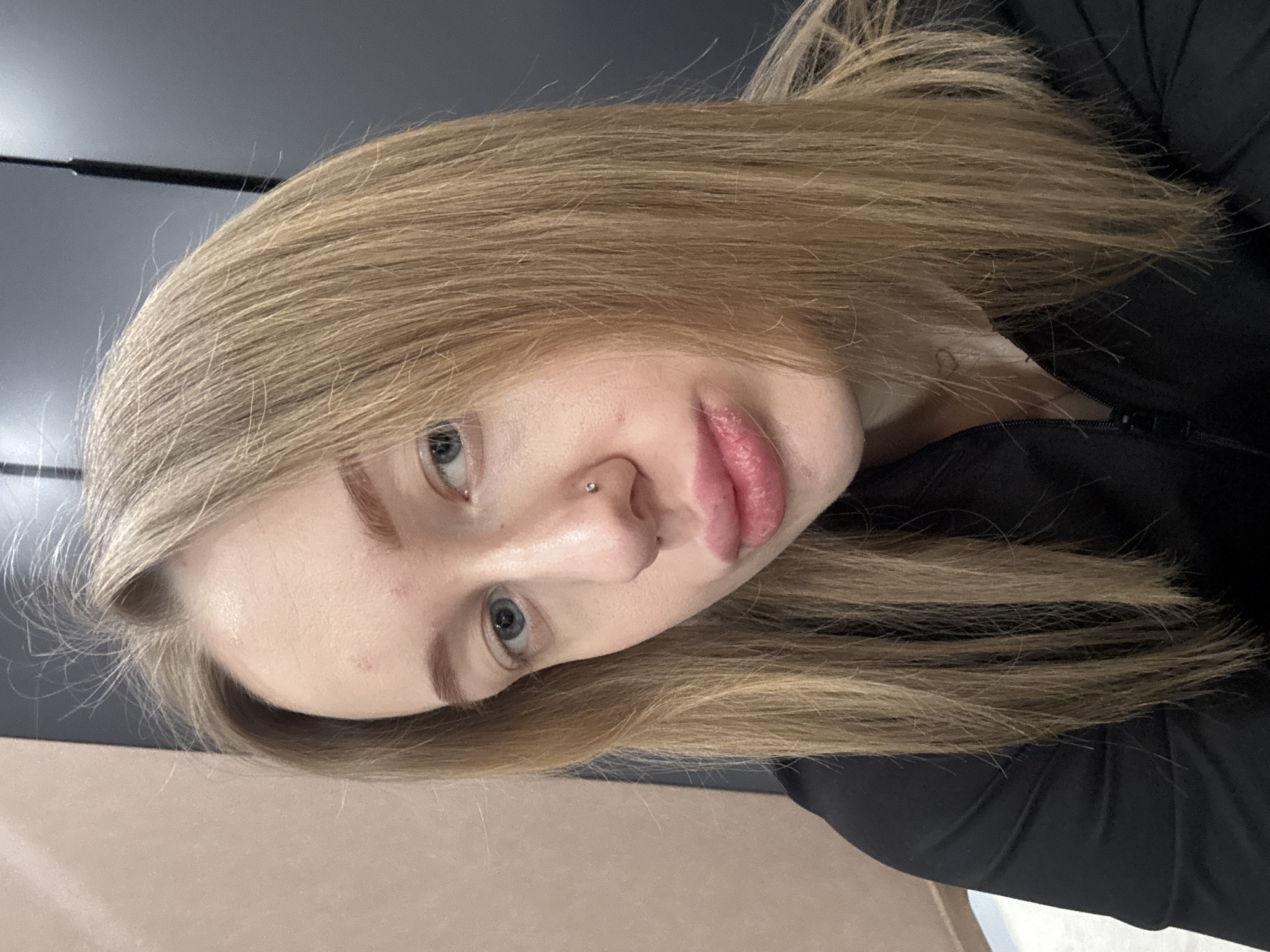 The image is a close-up selfie of a young woman with light blonde hair. She has fair skin, blue eyes, and a nose piercing. She is wearing a black top. There is a dark gray surface in the background, and a light brown surface is visible at the bottom of the frame. There is no information in the image that indicates it was taken in the city of Azov, Russia.