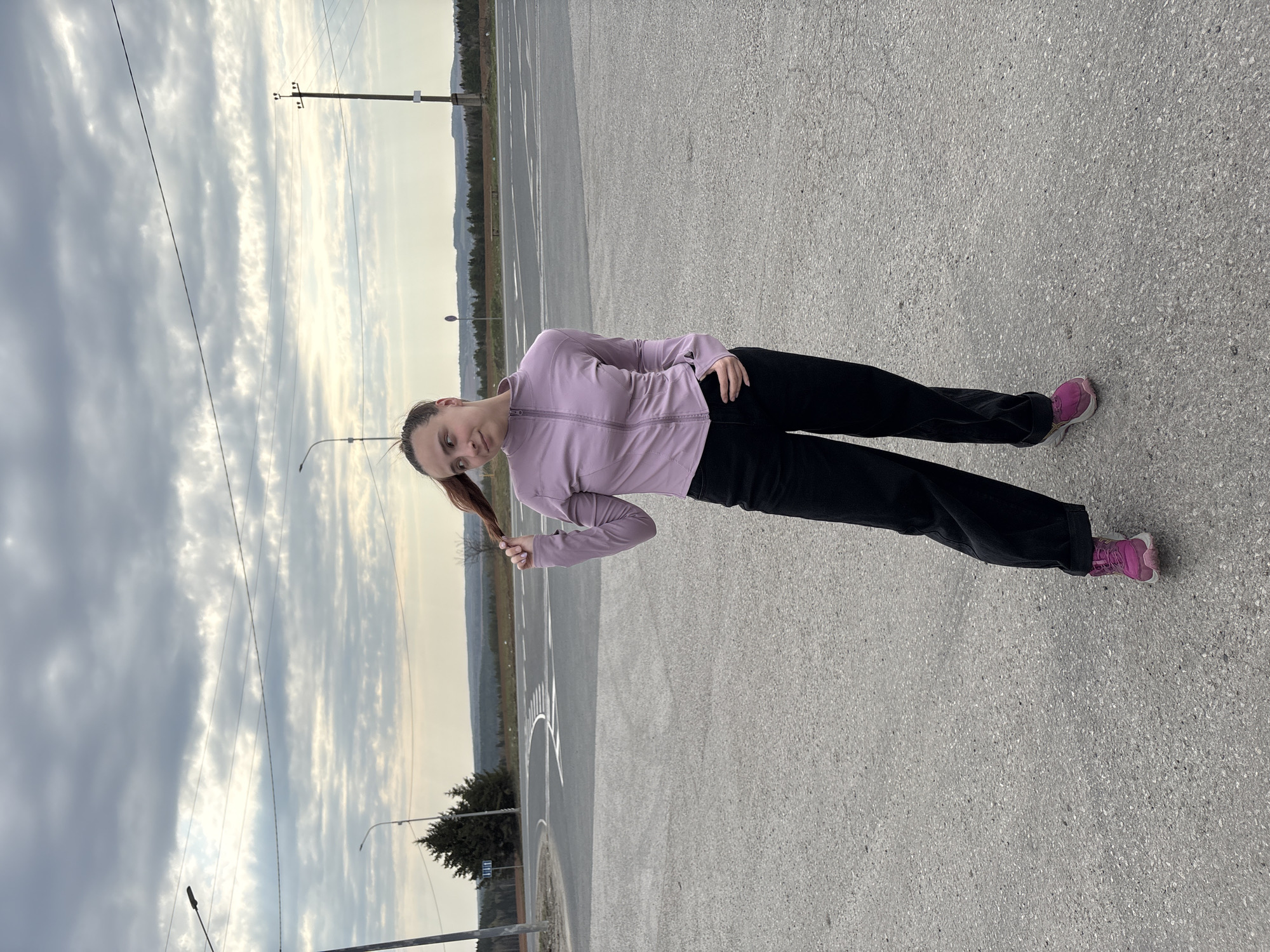 A young woman is standing in an open, paved area in Verkh-Sosnova, Russia. She is wearing a lavender jacket, black pants, and pink sneakers. She is holding the end of her ponytail in her left hand. The background includes utility poles, trees, and an overcast sky. White lines are painted on the pavement near where she is standing.