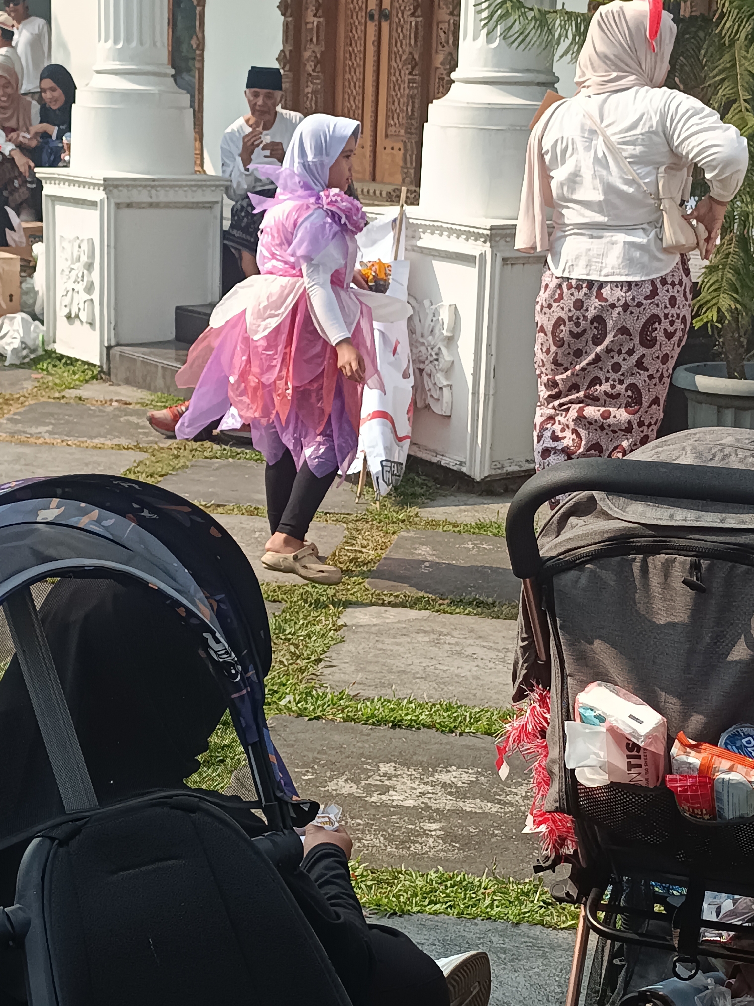 In Sleman, Indonesia, a young girl wearing a pink and purple layered dress with a flower embellishment around her neck and a light purple hijab is the central focus of this outdoor scene. She stands on a paved pathway bordered by grass, holding a small orange object. To her right, a woman in a light-colored shirt and a patterned skirt, also wearing a hijab, is seen from behind. In the foreground, the dark fabric of a stroller is visible, with a black hood and handles. A storage compartment on the stroller contains various items, including packaged wipes and small bags. In the background, an older man in a white shirt and black cap sits, appearing to eat or drink. Other individuals are visible in the background near a white pillar and an ornate wooden doorway. The scene suggests a gathering or event taking place outdoors, possibly during the day given the bright lighting.