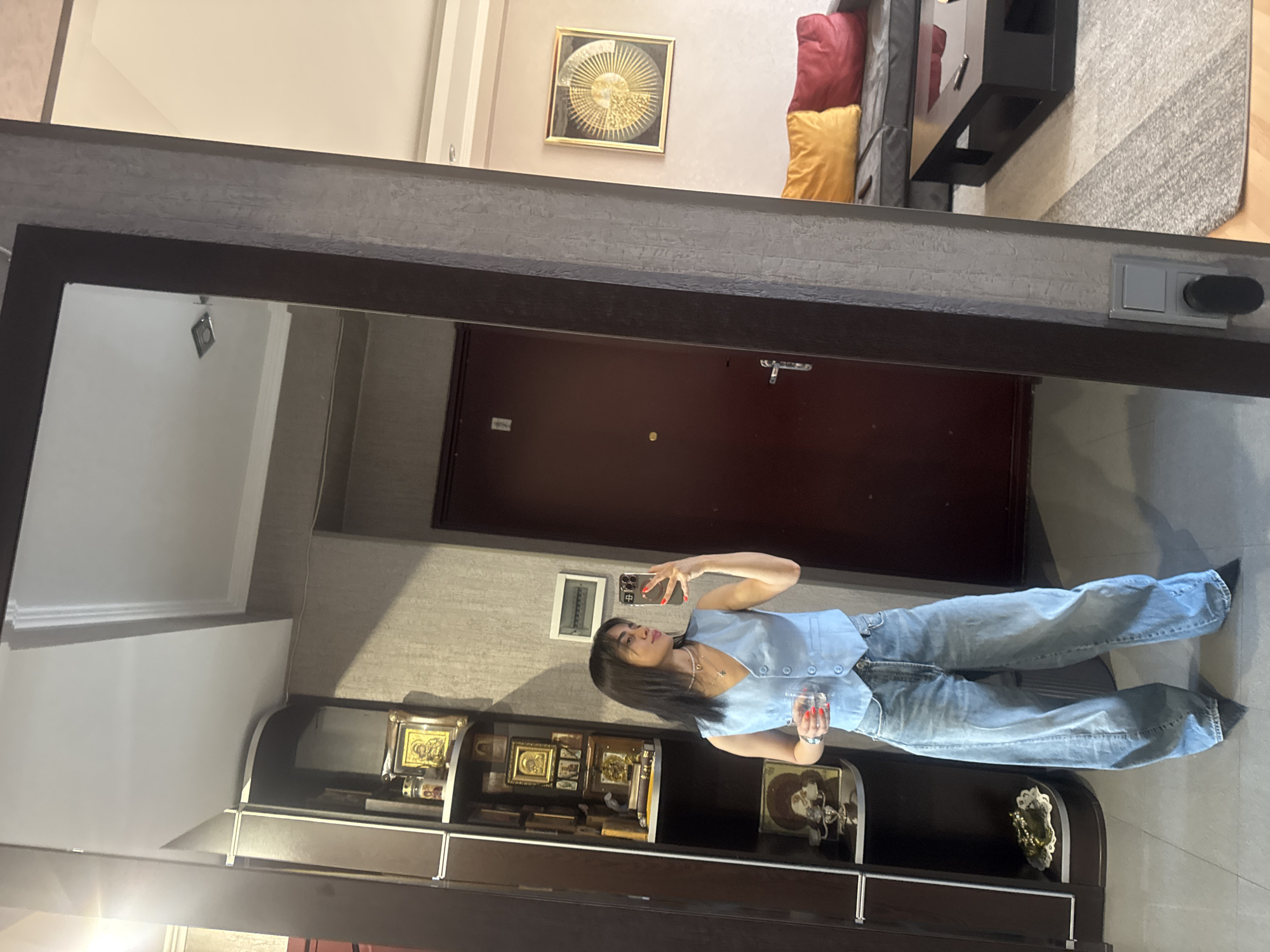 A woman is captured in a vertical, full-body mirror selfie, standing in an indoor hallway with a dark wood door and wall shelves displaying religious icons. She is wearing a light blue sleeveless vest and baggy light blue jeans, with a delicate pearl necklace around her neck. The woman holds a clear glass with a liquid in her right hand and her phone in her left, angled to take the photograph. Her dark hair is long and falls over her shoulders. The overall mood appears casual and self-aware. In the background, a glimpse of a living room can be seen with a modern sofa adorned with red and yellow cushions, and a black entertainment unit. The lighting suggests it is daytime, possibly afternoon, and the interior setting implies a residential or hotel environment in Tbilisi, Georgia.