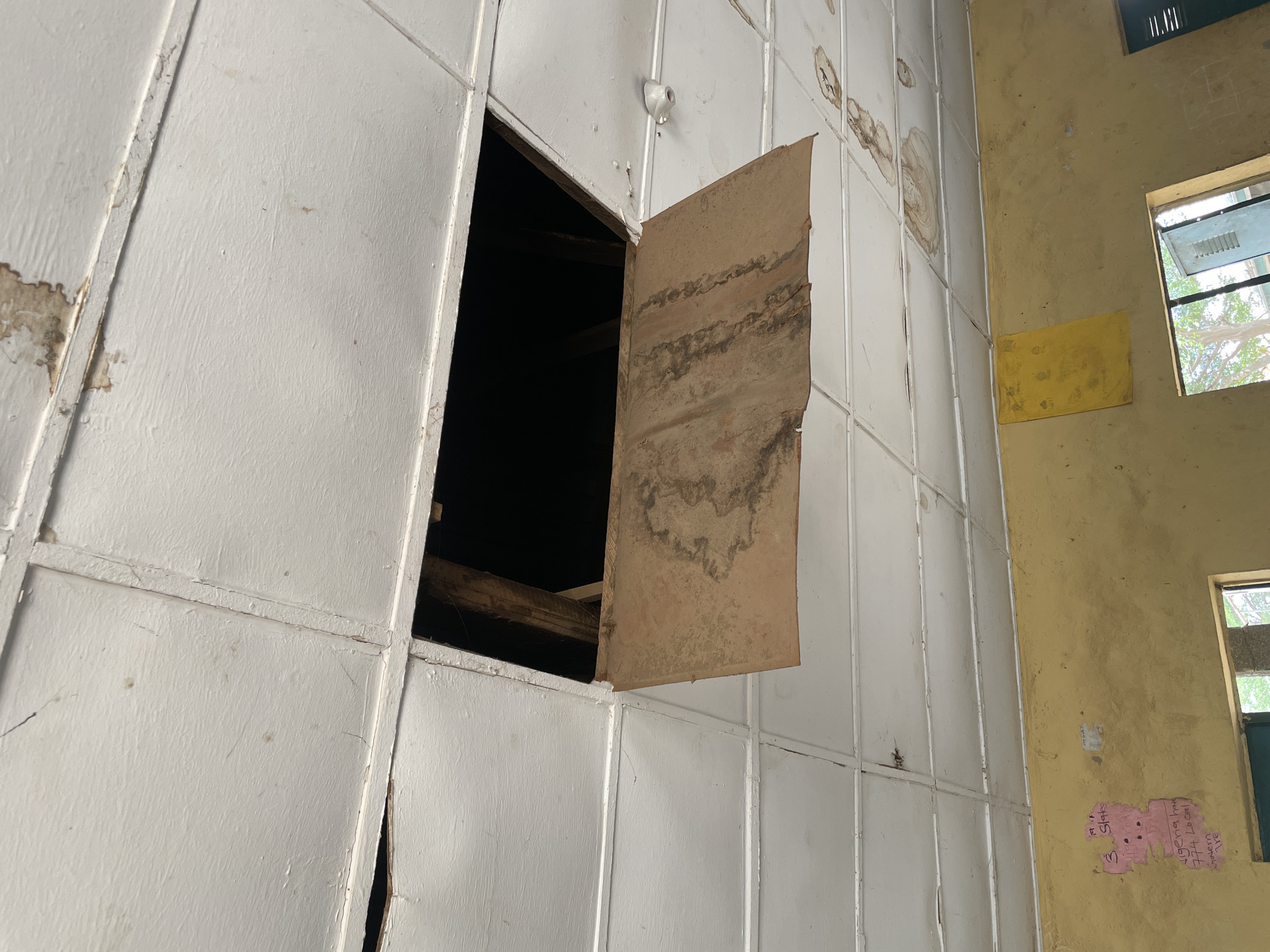 The image captures an indoor scene within an aged or neglected building, likely a classroom or public space, in Unguwar Rimi, Nigeria. The left and central portions of the frame are dominated by a wall composed of large, white, rectangular panels. Many of these panels show signs of significant wear, with peeling paint, cracks, and prominent circular dark stains indicating water damage.
A large, irregular rectangular opening is present in the upper-left section of the white paneled wall, revealing a dark, cavernous space behind it, with visible rough wooden beams. Adjacent to this opening, a detached, weathered brown panel hangs suspended. This panel, possibly made of fiberboard, is heavily stained with dark, wavy patterns, consistent with prolonged water exposure. Above the opening, a small, white ceramic electrical fixture, likely a lamp holder, is mounted on the white wall.
To the right, the wall is painted a distinct yellow color, which also shows signs of wear and staining. Two windows are set into this yellow wall; the upper one is partially open, revealing green foliage or trees outside, indicating it is daytime. Below the upper window, a plain yellow rectangular board or paper is affixed to the wall. Further down on the yellow wall, a torn piece of pink paper is visible, covered with handwritten text. The discernible text includes phrases such as "19 If States," "3," "Nigeria has," "774 Local," and "Government," suggesting educational or administrative notes related to Nigeria. The overall ambiance is one of disrepair and abandonment, hinting at a structure that has seen better days.