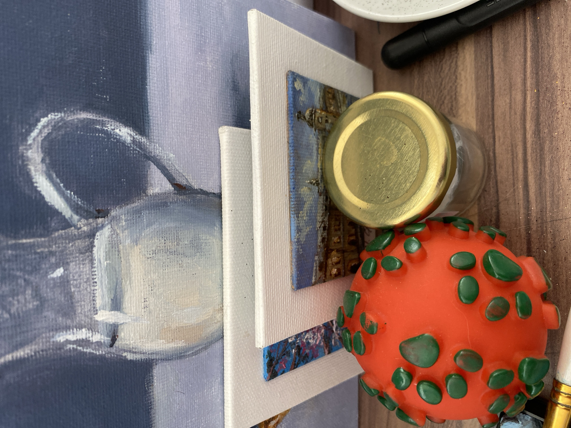 The image depicts a still life arrangement on a wooden surface. In the foreground, to the left, is an orange rubber toy with green protrusions, resembling a virus or spiky ball. To its right is a small, clear glass jar with a gold lid, partially filled with a white substance. To the right of the jar is a black marker pen and a portion of a white, speckled dish. In the background, several small canvases are stacked, displaying miniature paintings. The uppermost canvas shows a still life of a jug. The smaller canvases depict outdoor scenes, one with bare branches against a blue sky and another with a European-style building. The wooden surface appears to be a table. The lighting suggests an indoor setting, possibly a studio. The city of Bílina, Czechia is not depicted in this image.