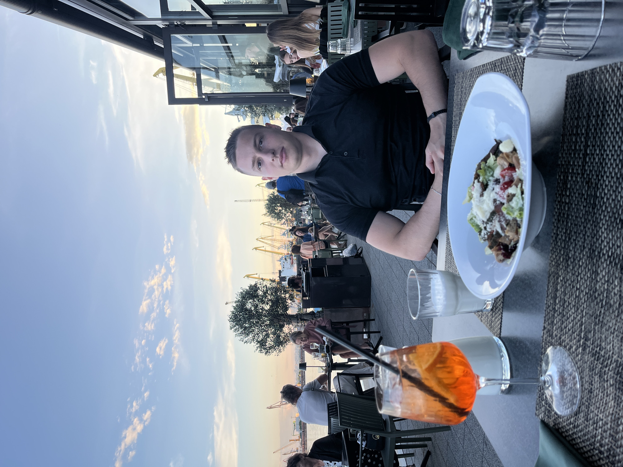 A young man in a black polo shirt sits at an outdoor table at a restaurant with a view of the sea and port. In front of him is a white bowl with a salad, an orange drink in a stemmed glass with a straw, and a glass of milk. He is looking directly at the camera. In the background, other diners are seated at tables. The sky is blue with scattered clouds. The setting appears to be the Port of Burgas in Burgas, Bulgaria, given the presence of cranes and ships in the background.