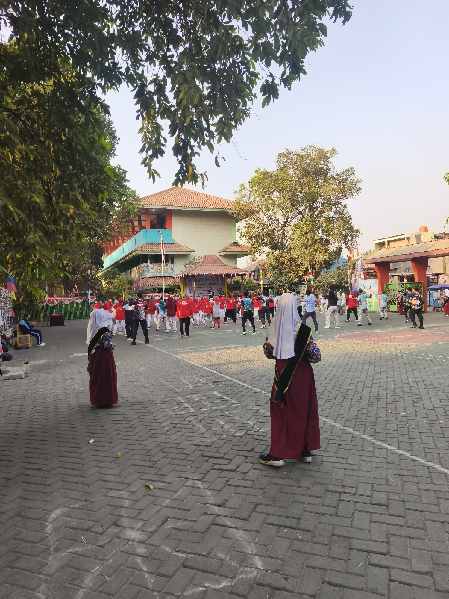 The image depicts an outdoor scene in North Jakarta, Indonesia, likely within a school compound, possibly during a morning exercise session. In the foreground, two individuals in traditional Islamic attire, a maroon robe and a white hijab, are facing away from the camera, observing a group of people engaged in synchronized movements. The group, predominantly dressed in red shirts, are spread across a paved courtyard, forming rows. The background features a building with a red-tiled roof and teal accents, likely a school building, adorned with Indonesian flags and banners. Trees with lush green foliage frame the scene, and the sky is a clear, bright blue. The ground is paved with interlocking bricks, and some white lines are visible, possibly marking exercise areas. Several other individuals are scattered around the periphery, some seated, others standing. The overall atmosphere appears to be one of communal activity and healthy lifestyle promotion.