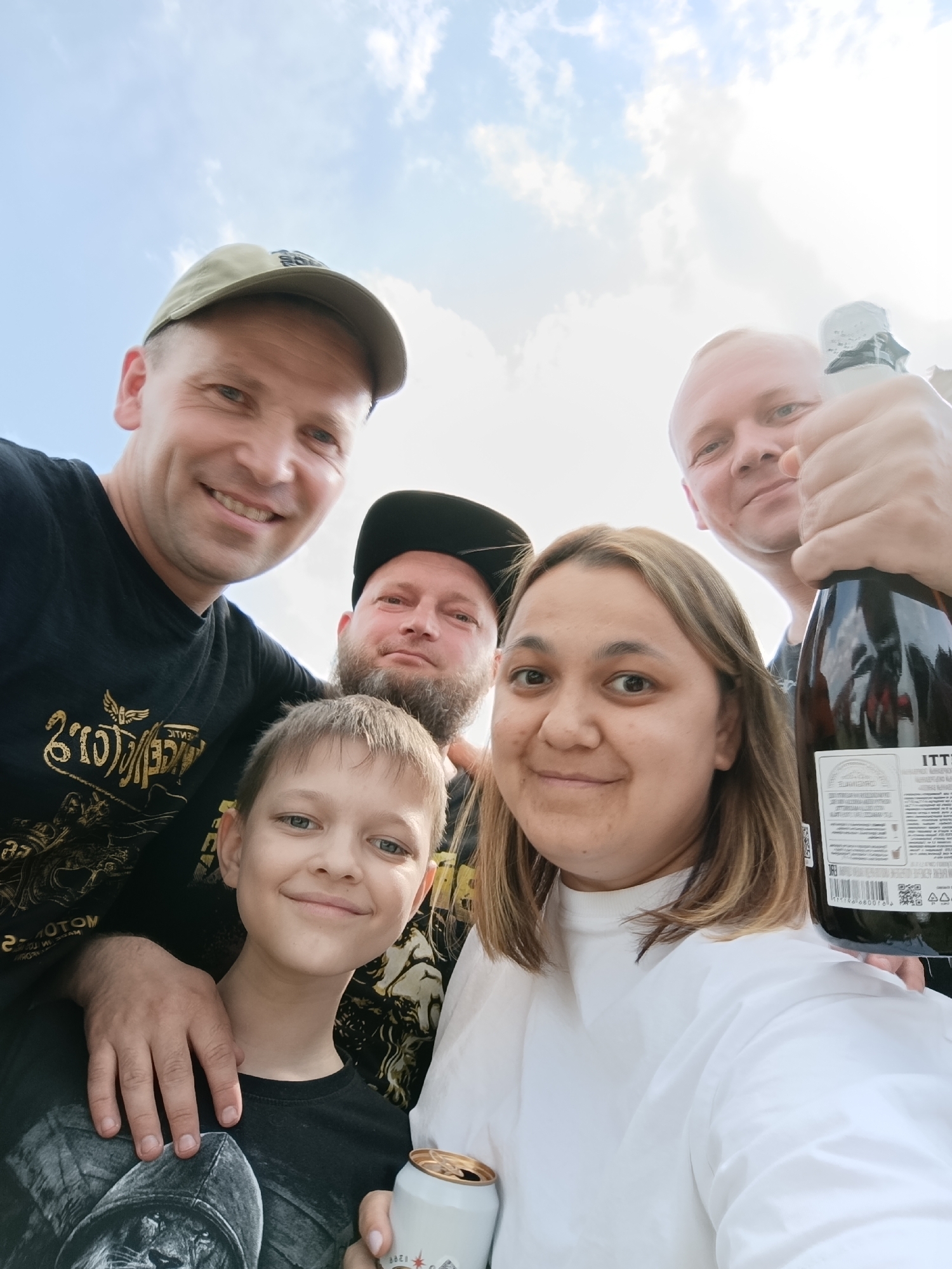 In a ground-level, wide-angle selfie, five people are visible, posing outdoors under a bright, partly cloudy sky, likely in or near the city of Lagunovo, Russia. The group consists of three men, one woman, and one young boy. The individuals are arranged in a somewhat staggered formation, with the boy and the woman in the front, and the men behind them.
The man on the far left, wearing a tan baseball cap and a black t-shirt with gold lettering, is smiling broadly at the camera. To his right, a young boy with short, light brown hair is also smiling. He is wearing a black t-shirt. Partially obscured behind the boy is a man with a beard, wearing a black baseball cap. To the right of the woman, who is wearing a white top and has brown hair, a man is holding up a bottle of champagne, smiling. The background consists of a light blue sky with scattered white clouds. The overall atmosphere suggests a casual gathering and celebration.